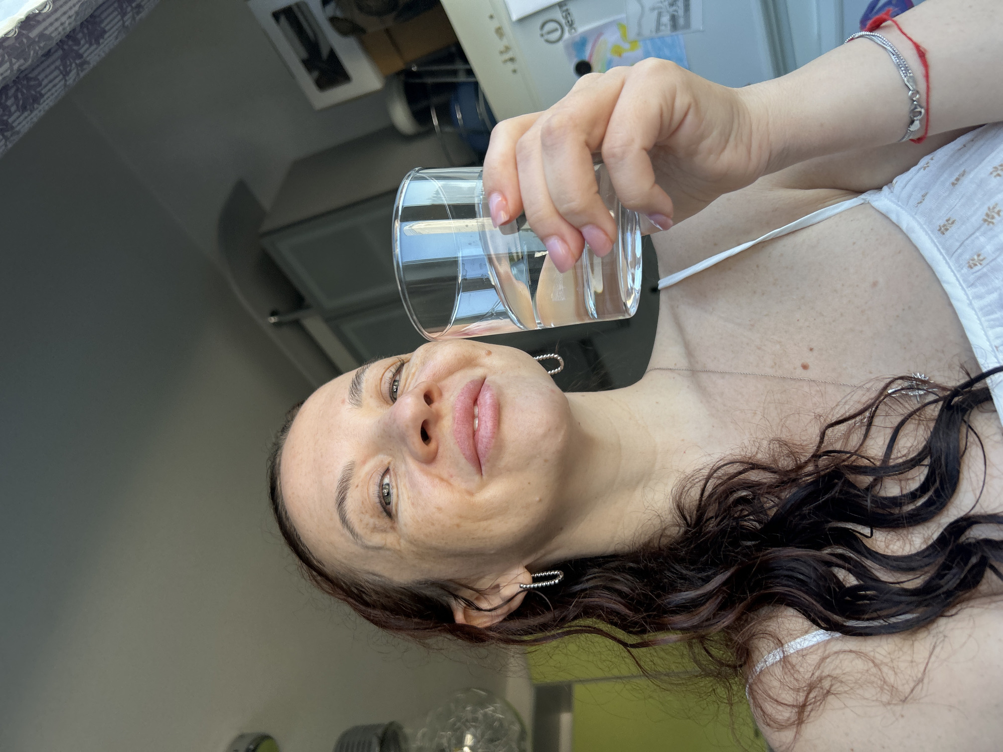 A woman is holding a glass of clear liquid in her right hand. She is wearing a white top, silver earrings, and a silver bracelet with a red string. Her hair is dark brown and curly. The background includes a kitchen area with cabinets and a white appliance. The image may have been taken in Chaikovsky, Russia.