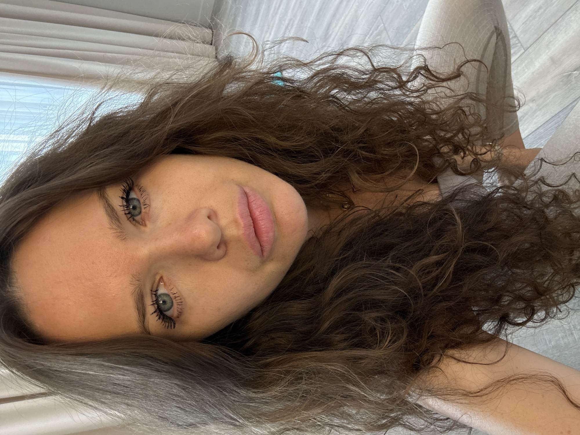 A young woman with long, wavy brown hair is pictured from the chest up, looking directly at the camera. She has blue eyes and her lips are slightly parted. She is wearing a light-colored top and possibly pants. The background shows light-colored curtains and a wooden floor. The image appears to have been taken indoors. There is no indication of the city of Khimki, Russia.