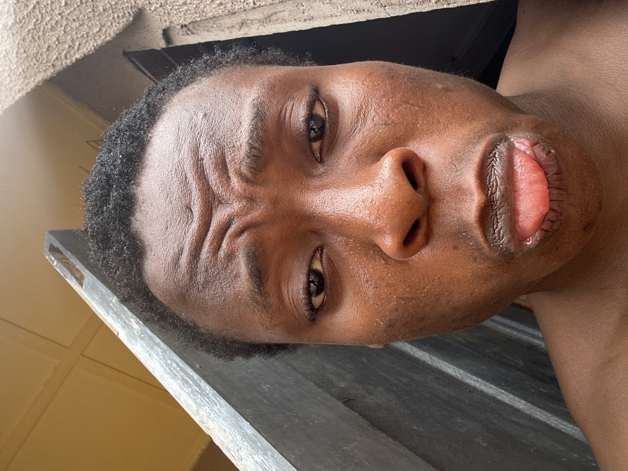 A close-up, angled view of a person's face. The individual has dark skin, short curly hair, and is looking directly at the viewer with furrowed brows. Their mouth is slightly open, revealing their tongue. The background is blurred, but there's a suggestion of a wooden structure or surface near the face. The city and specific location are not identifiable from this image.