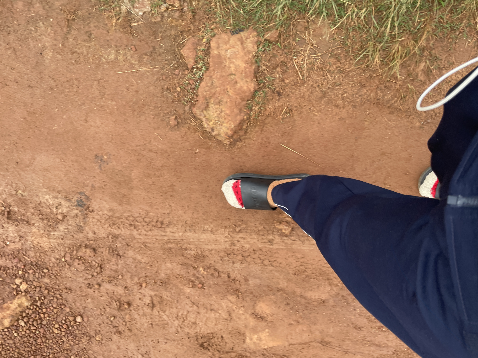 An overhead perspective shot captures a person's legs and feet walking on a dirt path. The ground is primarily reddish-brown soil, with scattered pebbles and patches of dry grass. A large, rough, reddish-brown rock is visible on the right side of the path. The person is wearing dark blue athletic pants and black slide sandals with red and white accents on the straps. A white cord, possibly from headphones, trails from the lower right corner of the frame. The scene appears to be in an outdoor, unpaved area, consistent with a rural or less developed part of a city like Durimi, Nigeria.