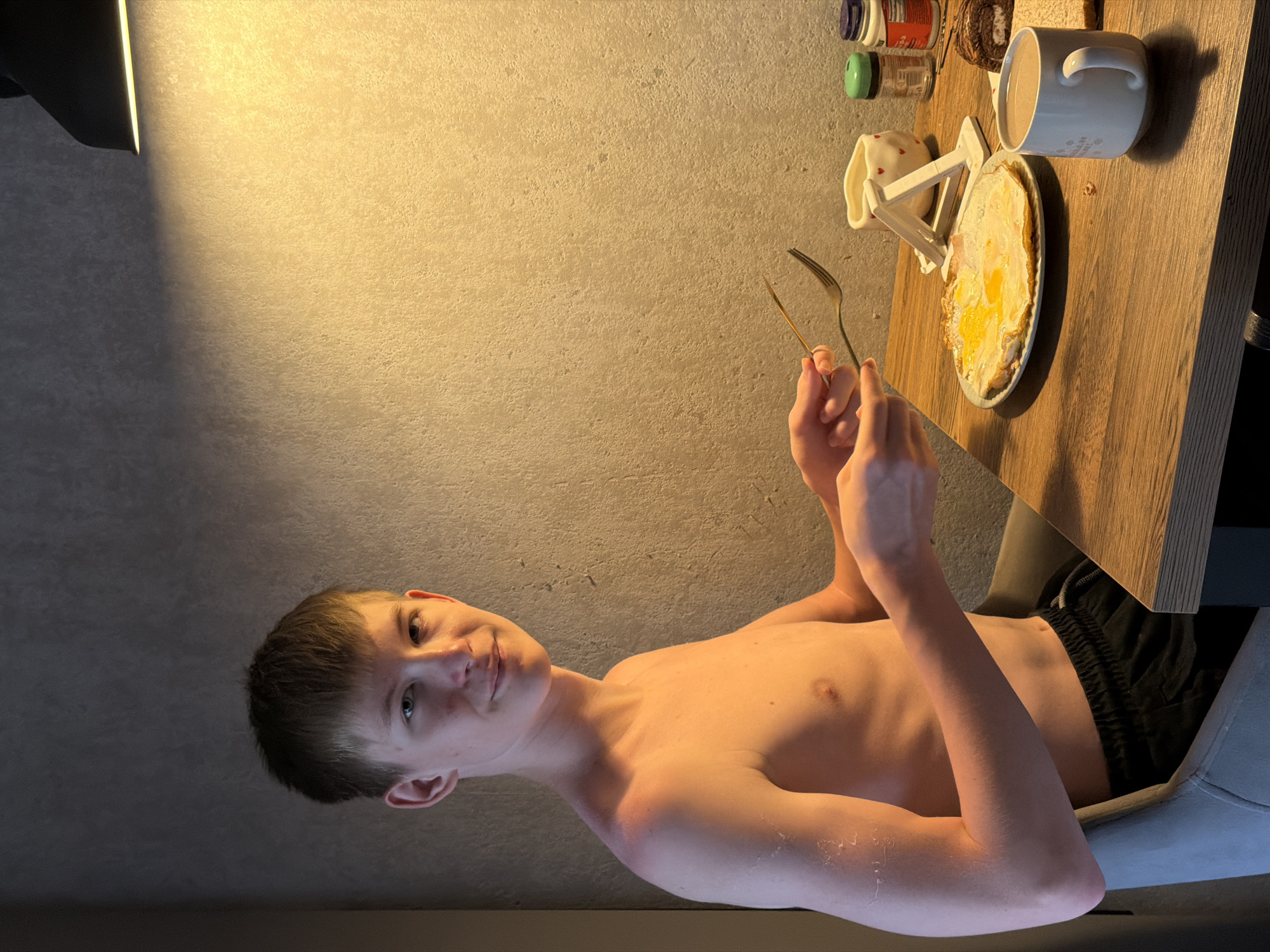 A young boy is seated at a wooden table, looking towards the camera with a slight smile. He is shirtless, wearing black shorts. The table is set with a plate of fried eggs, a white mug, a small white object with red hearts, and several small jars of spices or condiments. A fork and knife are held by the boy's hands. The setting appears to be a domestic interior, with a textured wall and a warm light source casting shadows from the left side of the frame. The time of day is suggested to be morning or early evening due to the warm lighting. The boy's expression is relaxed and friendly. The image is oriented vertically.