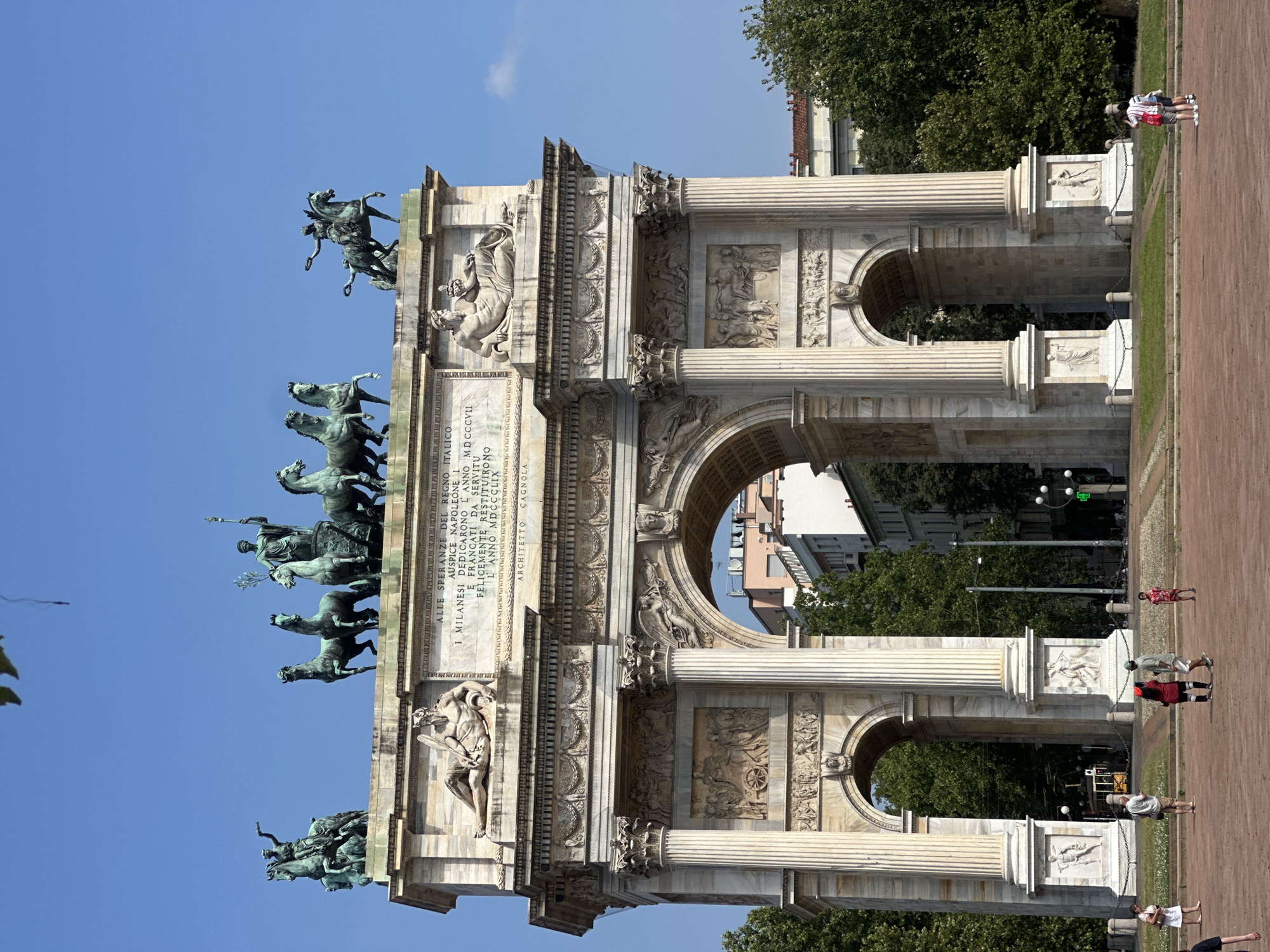 The image shows the Arco della Pace in Milan, Italy. The triumphal arch is made of white marble and is decorated with sculptures and reliefs. On top of the arch is a quadriga, a chariot drawn by four horses, with a statue of Victoria driving it. The arch has three passages, two smaller ones on the sides and a larger one in the center. The surrounding area is a park with green grass and trees. There are several people in the foreground, some walking and some standing. In the background, there are buildings and a tram. The sky is clear and blue. The overall scene is one of a sunny day in a public park with a prominent historical monument.