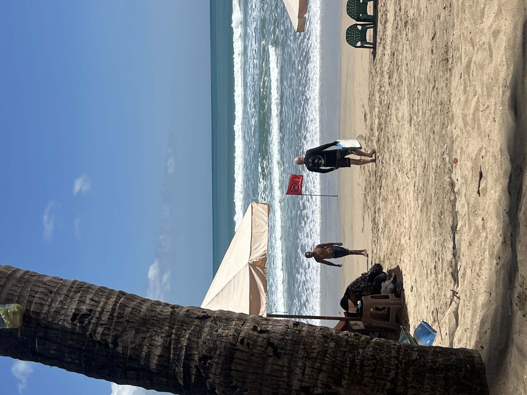A group of people are on a sandy beach with the ocean in the background. The sky is blue with some clouds. A palm tree is in the foreground, with its trunk partially visible on the left side of the image. There is a beach umbrella set up on the sand, providing shade. Two people are walking on the beach, one appears to be a man wearing a black long-sleeved shirt and shorts, carrying a white object. Another man is walking towards the ocean. In the distance, there are waves crashing on the shore. Two green plastic chairs are placed on the sand. A red flag is planted in the sand, indicating caution or a warning. This scene is characteristic of Kuta Beach in Kuta, Indonesia, a popular tourist destination known for its beaches and surf.