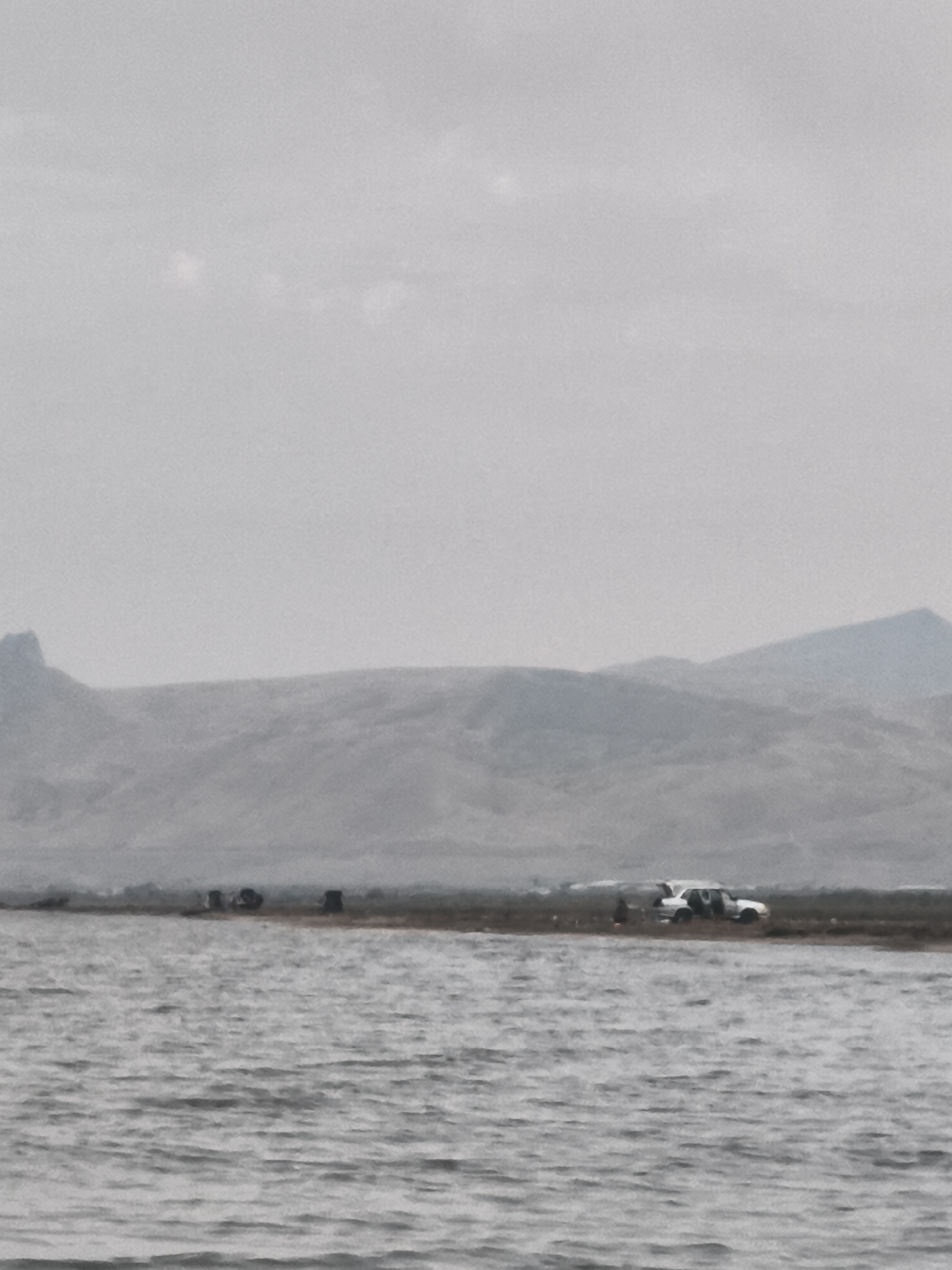 The image depicts a wide body of water in the foreground, with a hazy, overcast sky above. In the middle ground, a narrow strip of land is visible, characterized by a sparsely vegetated, dry terrain. Along this land strip, several dark, upright objects, possibly benches or small structures, are scattered. Further to the right on the land, a white vehicle, possibly an SUV or van with its doors open, is parked. Another dark object is near the vehicle, and what appear to be smaller dark objects or figures are visible behind the main group of dark upright objects. In the background, rolling hills and mountains create a layered, muted landscape under the diffused light. The overall scene suggests an outdoor location, potentially a natural or recreational area near Balaca Həmyə, Azerbaijan.