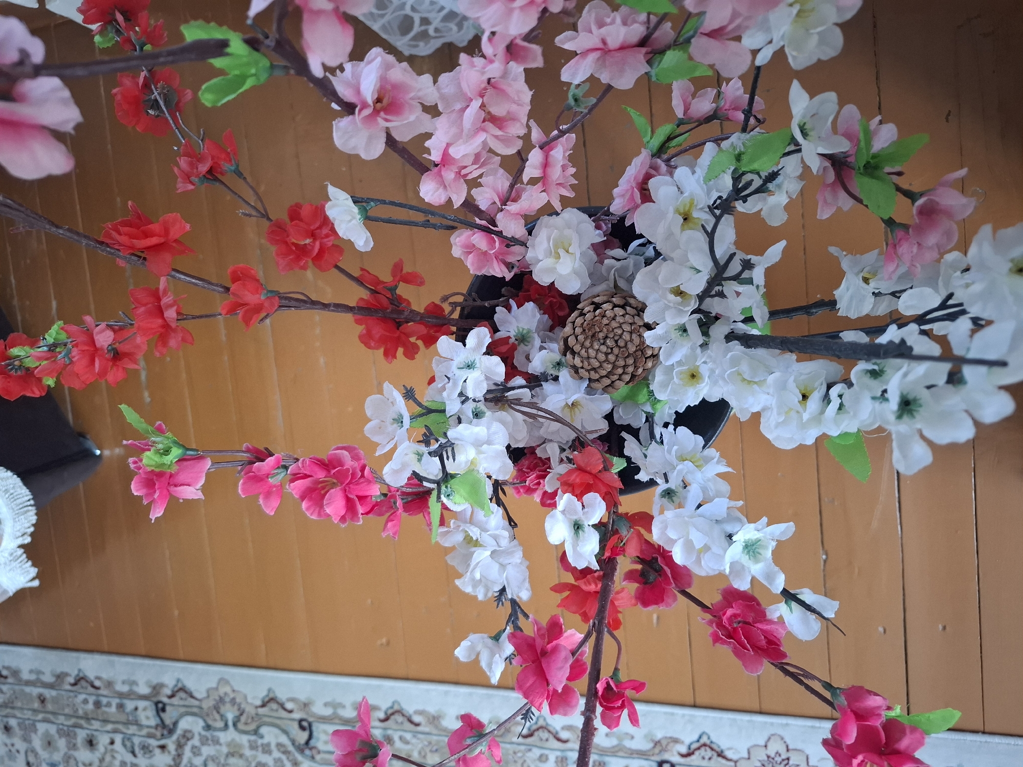 A top-down, close-up shot shows a faux floral arrangement with pink, white, and red flowers and green leaves, presented in a black vase, situated on a wooden floor. The background features a portion of an oriental-style rug on the left and a white lace curtain to the right. No people are visible. The setting appears to be an indoor environment, likely a residential room, within Almaty, Kazakhstan.