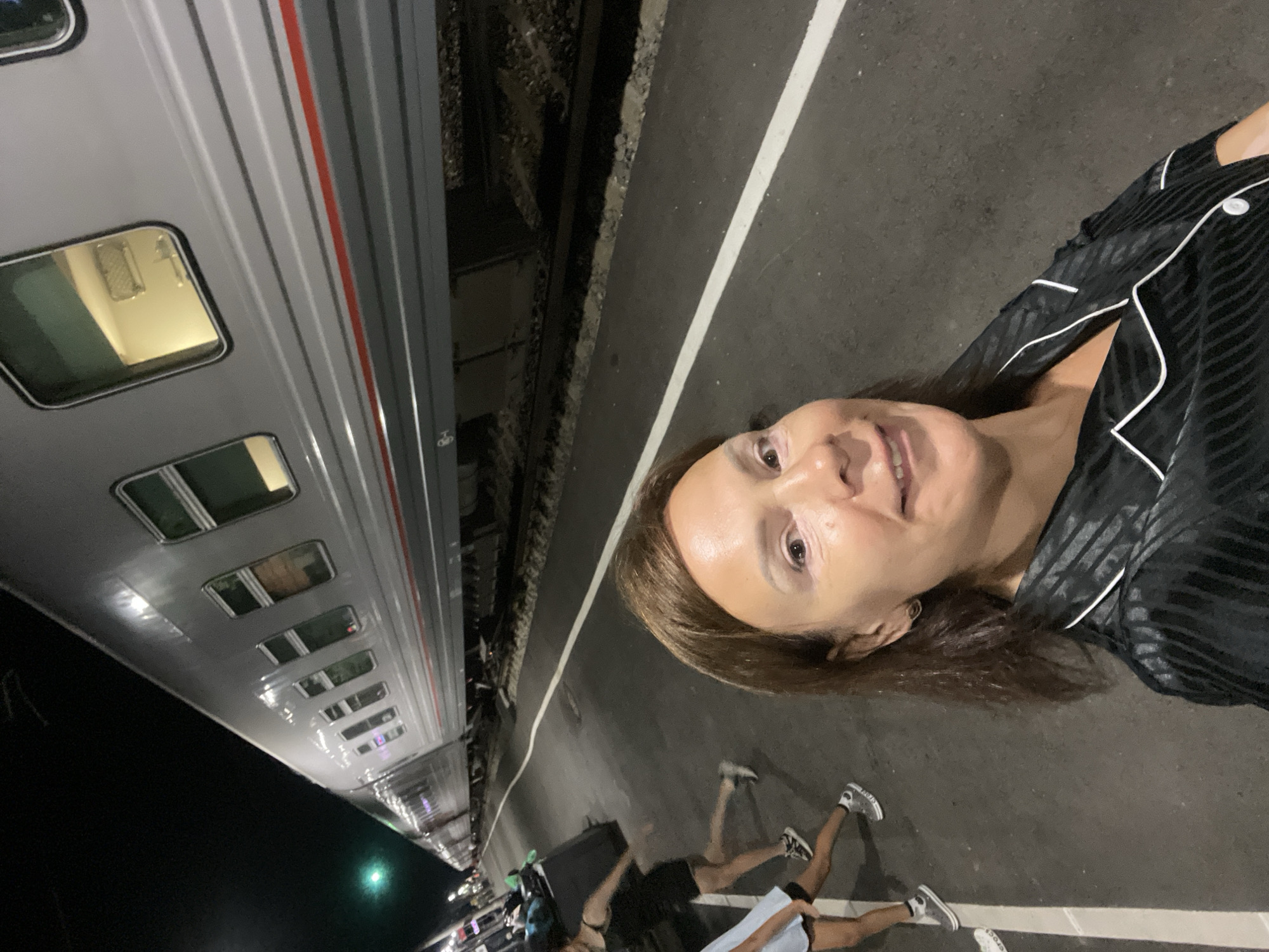 A night-time selfie is taken by a woman standing on a train platform in Goryachiy Klyuch, Russia. The woman, with brown hair and wearing a black striped pajama top, smiles at the camera. Behind her, a long, gray passenger train is stopped at the platform, its windows illuminated from within. The platform itself is dark asphalt with a white line marking the edge. In the background, two other individuals, one in a black dress and another in a light blue top, are walking away from the camera. Luggage is visible near them. The overall scene suggests travel or an arrival/departure at the train station.