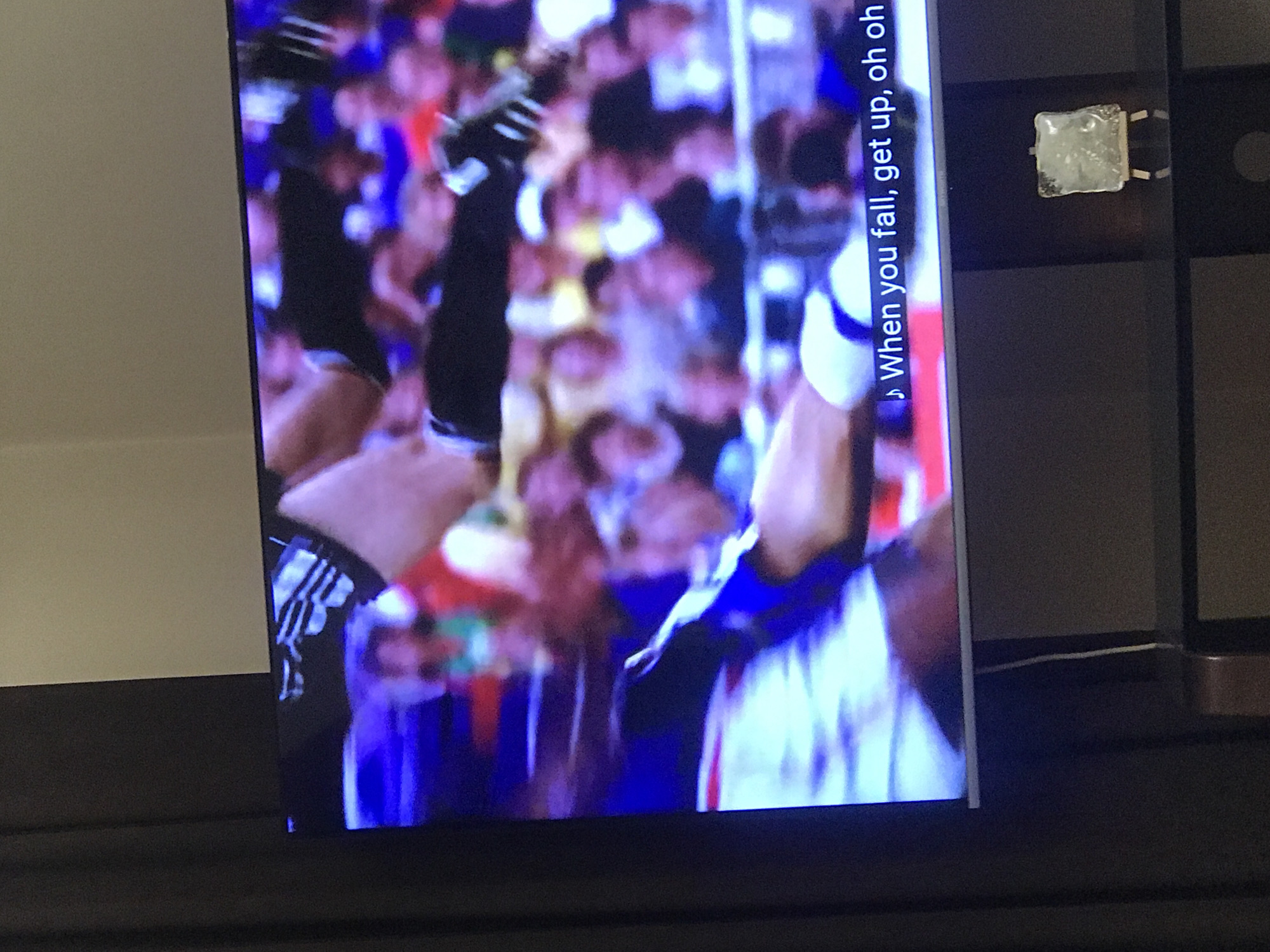 A photo shows a close-up shot of a television screen, with the picture displaying a crowded stadium or venue where people appear to be celebrating or cheering. Hands are raised in the air, some wearing gloves or wristbands, and the crowd is blurred in the background. Subtitles on the right side of the screen read "When you fall, get up, oh oh oh." The TV sits on a dark surface, which is likely a piece of furniture. Beside the TV, a dark-colored shelving unit or cabinet is visible. There is no information provided about this image being taken in Yerevan, Armenia.