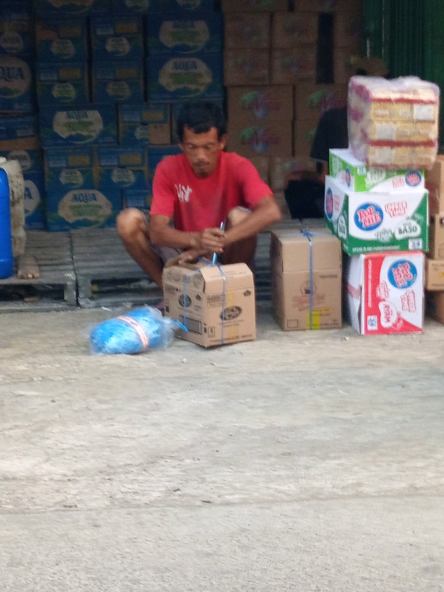 A man in a red shirt is crouched down next to a stack of cardboard boxes on a concrete surface. The boxes are various sizes and some have labels with writing and logos. To the man's left, a blue plastic-wrapped object, possibly a water jug, lies on the ground. In the background, more stacked boxes are visible, many labeled with "AQUA". The environment appears to be an outdoor area, possibly a market or a loading dock. The lighting suggests it is daytime. The presence of "AQUA" branding strongly indicates the location is Karawang, Indonesia, a major center for Aqua bottled water production.