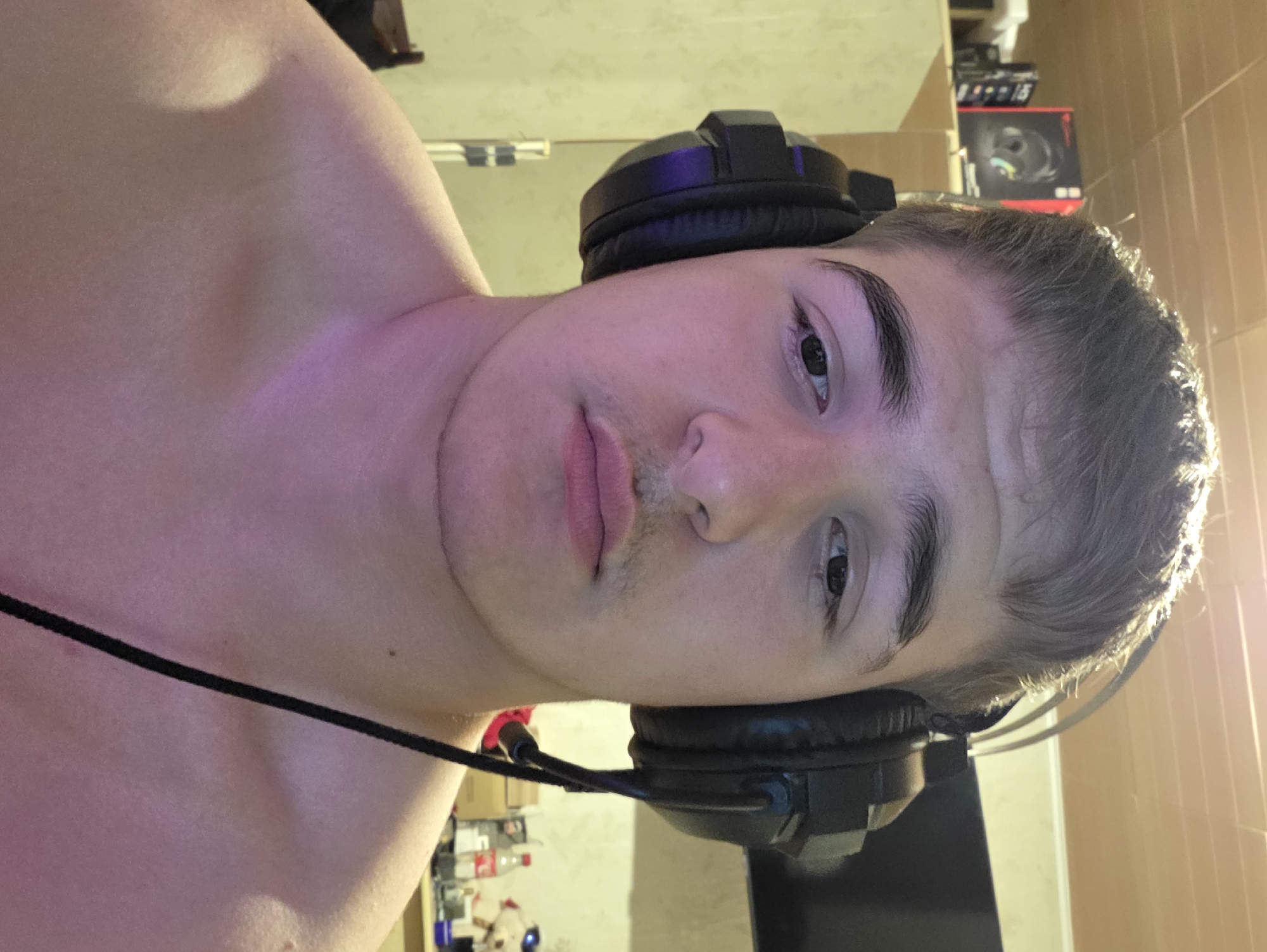 A young man with short brown hair and headphones over his ears is facing the camera. His eyes are open and he is looking directly at the camera with a neutral expression. He has a light mustache. His shoulders and upper chest are visible, and he is not wearing a shirt. The background consists of a wall with a tiled ceiling, shelves with boxes and other items, and a dark screen in the upper right corner. The lighting in the room is warm and casts a pinkish hue on the man's skin. The overall scene appears to be a casual indoor setting, possibly a bedroom or gaming area, in the city of Aleksandrovo, Bulgaria.