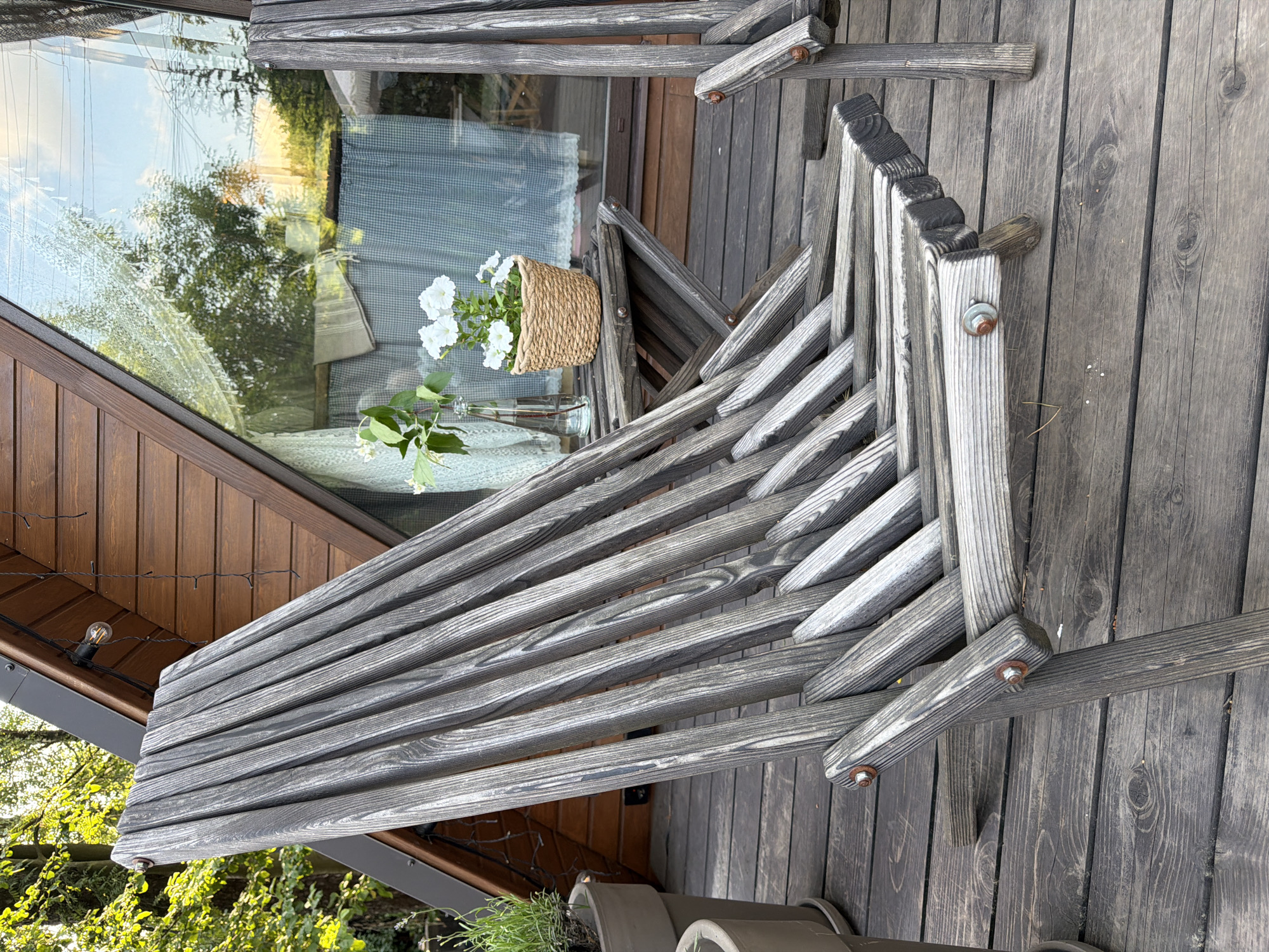 The image shows a weathered wooden deck outside an A-frame cabin. A grey wooden lounge chair is in the foreground, angled towards the right. Behind the chair, a small wooden table holds a woven basket with white flowers and a glass bottle with greenery. A large triangular window with a checkered curtain is visible on the cabin wall. Trees and a cloudy sky are reflected in the window. The deck is made of dark, weathered wooden planks. The overall scene suggests an outdoor seating area in a rustic, natural setting.