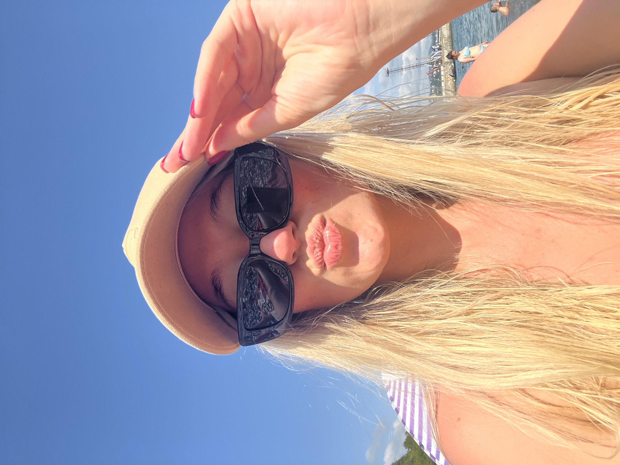 A blonde woman wearing a tan baseball cap and black sunglasses is taking a selfie outdoors on a sunny day. She is puckering her lips and touching the brim of her cap with her right hand, which has red nail polish. Her long blonde hair is wet and falls around her shoulders. In the background, there is a blue sky, the sea, a striped beach chair, and a few people swimming in the water. The scene appears to be a beach or waterfront area in the city of Tuapse, Russia.