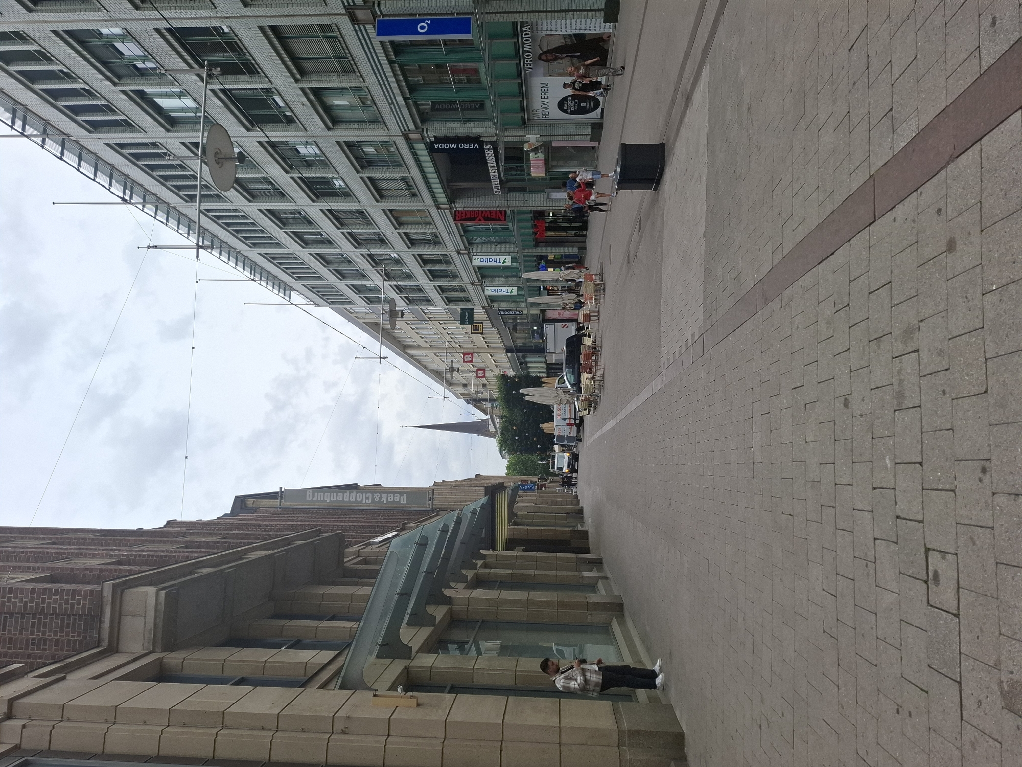 The image shows a street in Hamburg, Germany, likely a pedestrian shopping area. The left side of the street features a building with a brick upper story and stone facade. A person stands near the entrance of a store on this side. The right side of the street is lined with a modern, multi-story building with many windows. Several shops are visible along this side, with awnings and signage. There are streetlights and utility wires overhead.
In the background, a church spire is visible. Further down the street, there are several vans and a few parked cars. Some outdoor seating areas with tables and chairs are set up on the right side of the street. A few people are walking along the street. The ground is paved with interlocking rectangular stones. The sky is overcast.