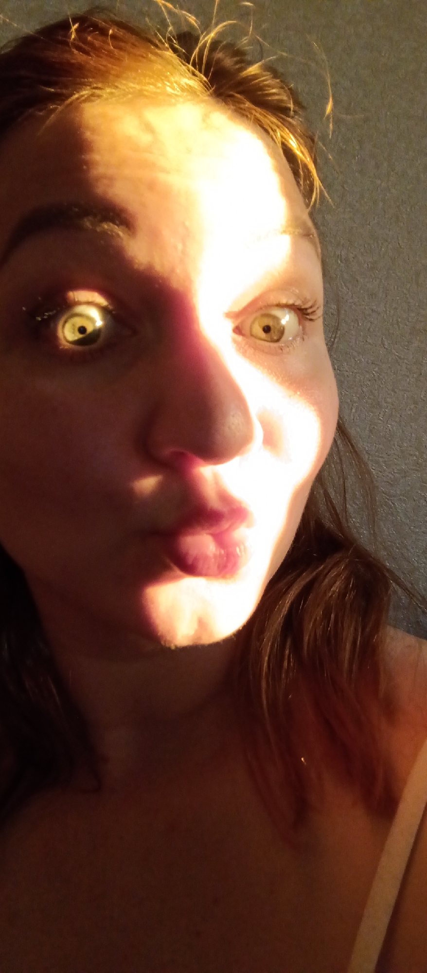 A close-up portrait of a fair-skinned woman with light brown hair. Harsh, direct sunlight illuminates the left side of her face, casting strong shadows on the right. She has a pursed lip expression, possibly posing for a "duck face." Her eyes are light-colored, with one appearing yellow due to the sunlight. The background is a blurry gray wall. The location is unknown.