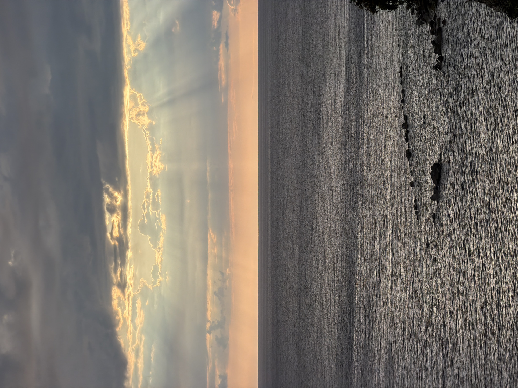 This media file captures a dramatic sunset or sunrise over the sea near Mirište, Montenegro. The image is composed of two distinct halves separated by a clear, straight horizon line.
The upper half features a captivating sky, dominated by a thick bank of clouds. The sun, obscured by these clouds, casts brilliant golden and orange light from behind, creating prominent crepuscular rays that pierce through gaps in the cloud cover. The clouds themselves range in color from dark grey where shadowed to glowing gold and orange where backlit by the sun.
The lower half of the image displays the vast expanse of the sea. The water surface is textured with countless small ripples and glints, reflecting the low-angle light and appearing as a shimmering, silvery-grey tapestry. In the bottom right corner, a dark, rocky coastline or cliff edge is visible, with a series of individual rocks extending from it into the textured water, creating a stark silhouette against the shimmering surface.
The overall scene conveys a tranquil yet awe-inspiring natural moment, likely at dusk or dawn, given the warm, dramatic lighting. There are no people, explicit activities, or visible text in the image.