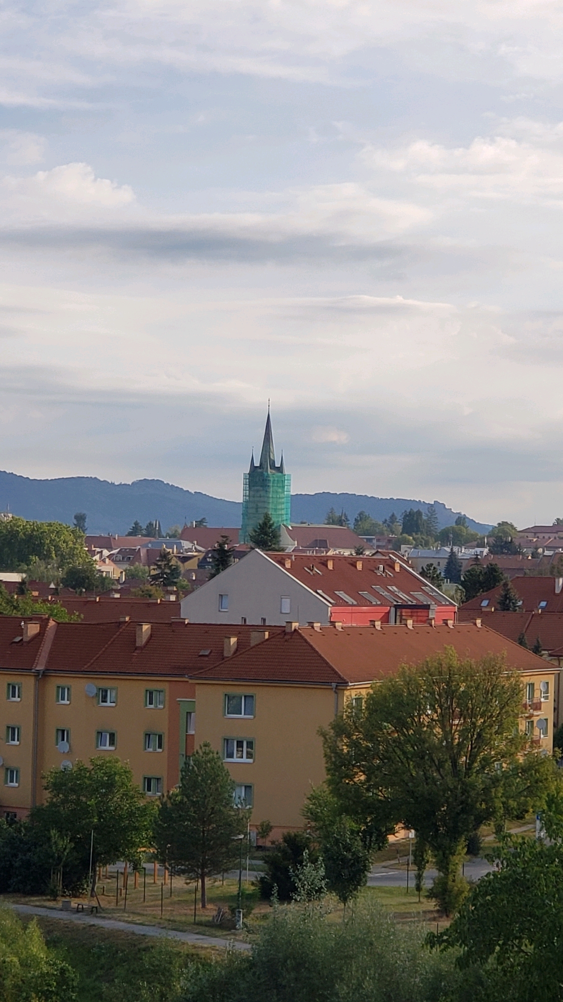 This is a vertical, slightly elevated, medium-range shot capturing a cityscape in Prešov, Slovakia. The image is dominated by residential buildings with orange-tiled roofs. In the midground, a tall green church steeple, partially covered in scaffolding, rises above the rooftops. Behind the buildings, rolling hills are visible under a pale blue sky with scattered white and gray clouds. In the foreground, a park-like area with trees and some playground equipment is visible. The overall scene depicts a suburban or residential area within the city, likely on a pleasant day. There are no people visible in the image.