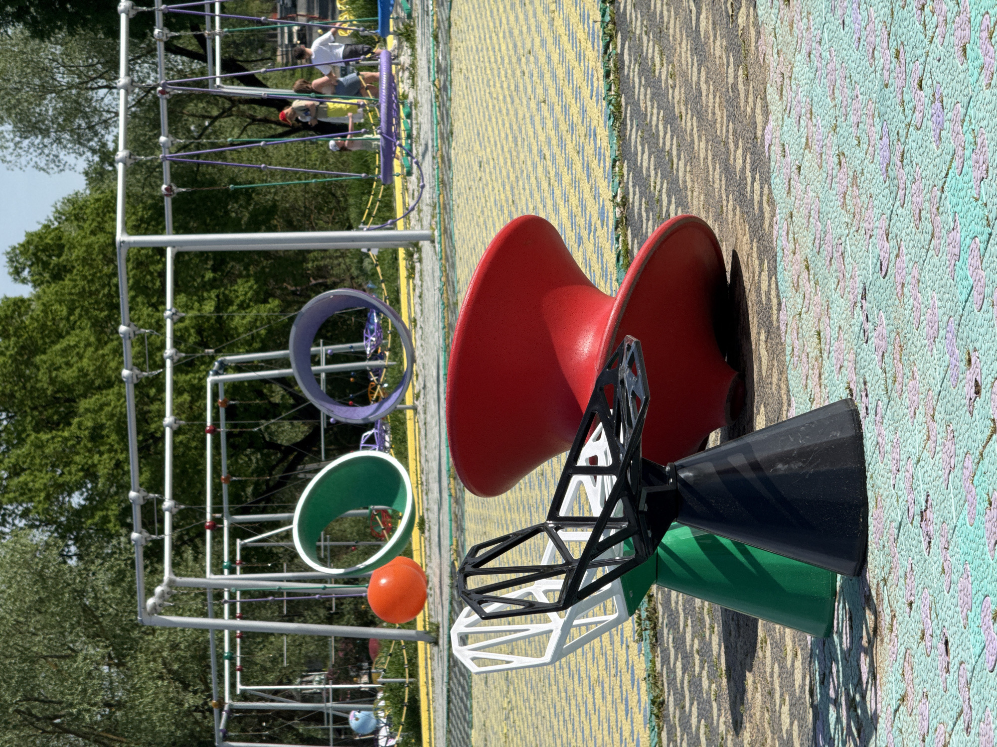 This image shows a playground in the city of Синьково, Russia. The playground features a complex climbing structure with interconnected platforms and rope bridges in the background, where a few people are visible. In the foreground, there's a colorful geometric play structure with red, black, green, and white components. An orange ball hangs on the edge of the climbing structure. The ground is covered with patterned safety mats. The area is surrounded by trees.