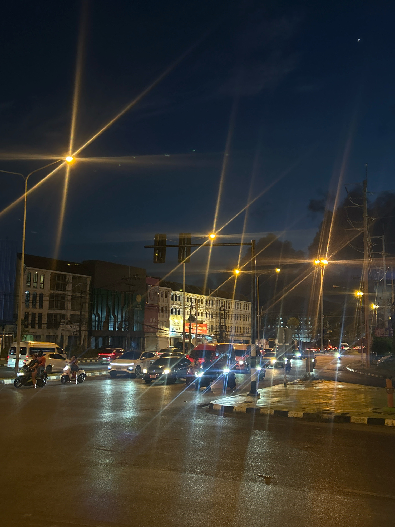 The image depicts a street scene at dusk or night in Ratsada, Thailand. The road surface appears wet, reflecting the light from streetlamps and vehicle headlights. Multiple streetlights with prominent starburst flares are visible throughout the frame, illuminating the dark sky and the surrounding urban environment. Buildings line the street on the left, displaying a mix of architectural styles, some with illuminated windows. Traffic is present, including cars and motorcycles. On the left side of the road, two individuals are riding a motorcycle, and another motorcycle is parked nearby. Further down the road, several cars are driving, their headlights on. On the right side of the image, utility poles with visible wiring are present. The overall impression is a busy urban intersection at night with moderate traffic and clear visibility due to artificial lighting.