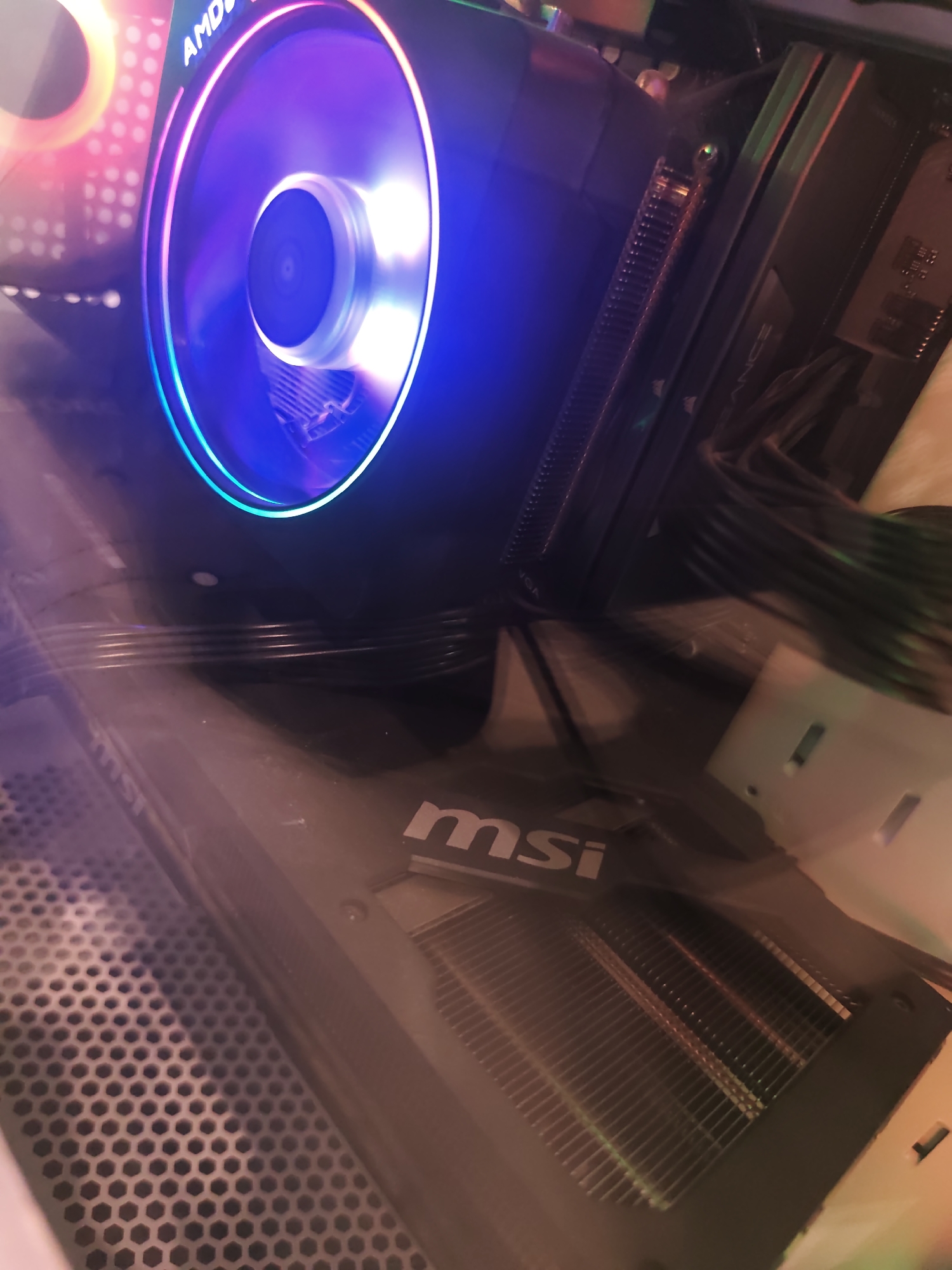 The image displays the illuminated interior of a desktop computer case, viewed at an angle through a reflective side panel. Dominating the upper center is an AMD CPU cooler, featuring a circular RGB fan that emanates vibrant blue, purple, and hints of pink and red light. The "AMD" logo is partially visible along the top edge of the cooler. To the right of the CPU cooler, several RAM modules are visible, with "CORSAIR VENGEANCE" branding partially discernible on one stick. Below the CPU cooler and extending into the lower half of the frame is a graphics card, identified by the prominent "msi" logo on its side and visible cooling fins beneath it. Various black cables are routed near the RAM and behind the graphics card. On the lower left, a grey, hexagonal perforated metal panel, likely for airflow, is visible. Another internal RGB fan or light source with red and orange hues is partially visible in the upper left background. The overall scene is characterized by the colorful internal lighting of the components.