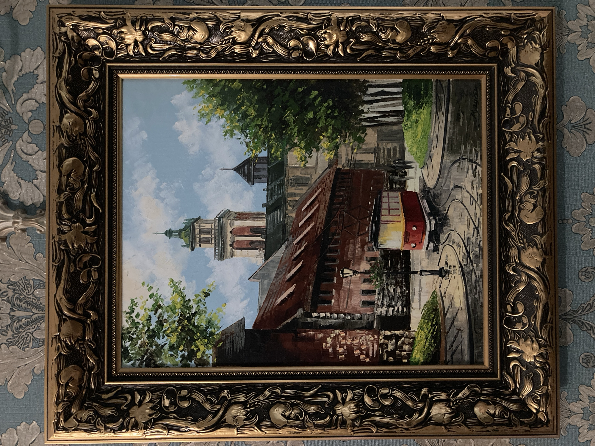 The painting depicts a street scene in what appears to be an older European city. In the foreground, a red tram with a yellow stripe is stopped on wet cobblestone tracks. To the left of the tram is a weathered brick building with a reddish-brown roof, and to its right, a patch of green grass and several slender trees. In the background, a multi-story building with a prominent tower topped by a green dome is visible, suggesting a church or a significant historical structure. The sky is blue with white clouds. The painting is enclosed in an ornate, antique gold-colored frame with intricate carvings. The overall impression is that of a charming, possibly historic, urban setting on a damp day.