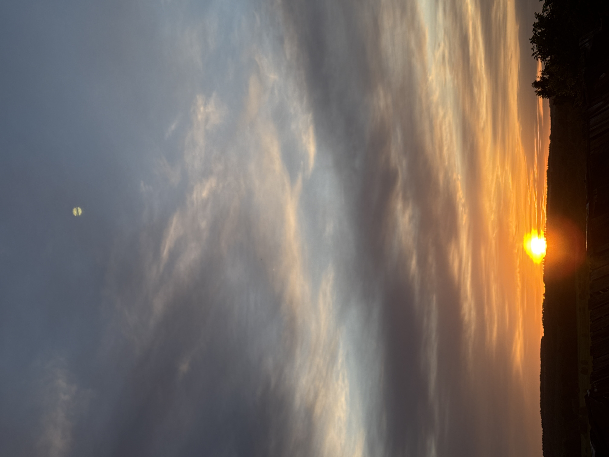 The image depicts a sunset in Nizhnyaya Gar', Russia. The sun is visible on the right side, partially obscured by the horizon. The sky is filled with streaks of light, transitioning from golden hues near the sun to grayer shades on the left. A small, light-colored object is visible in the gray area of the sky on the left. The horizon line includes a dark, silhouetted area of trees or vegetation.