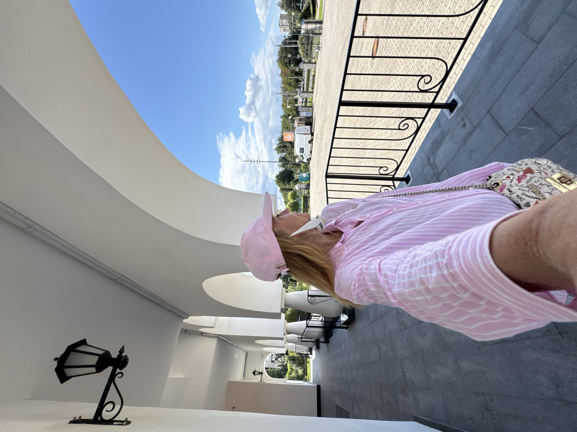 A woman wearing a pink baseball cap and a pink and white striped button-down shirt stands in front of a white arched walkway in Saint Petersburg, Russia. She is holding a patterned handbag with a gold clasp and chain strap. Her face is turned to the right, and she appears to be looking out at a plaza with a black metal railing in the foreground. In the background, there are trees, buildings, and a white van. The sky is blue with scattered clouds.