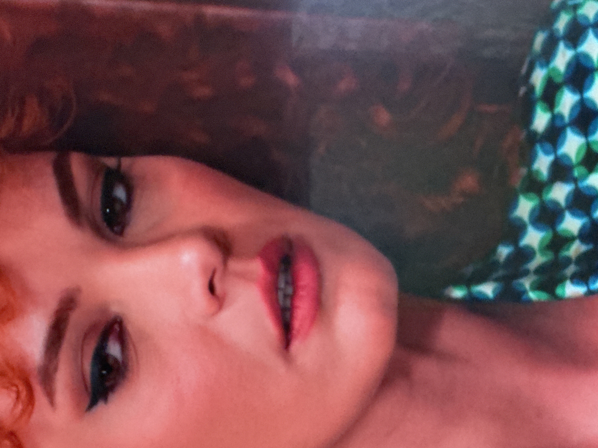 The image is a close-up portrait of a woman, focusing on the left side of her face. She has auburn hair styled in loose curls, and her skin is illuminated by a warm, pinkish light. Her left eye is visible, adorned with dark eyeliner and mascara, and her eyebrow is neatly shaped. The woman is wearing a patterned garment, predominantly green and blue, which is partially visible in the lower right portion of the image. The background is dark and out of focus, with hints of reddish-brown tones, suggesting an indoor setting. The provided location of Uyutnoye, Ukraine, is not discernible from the visual information in the image itself.