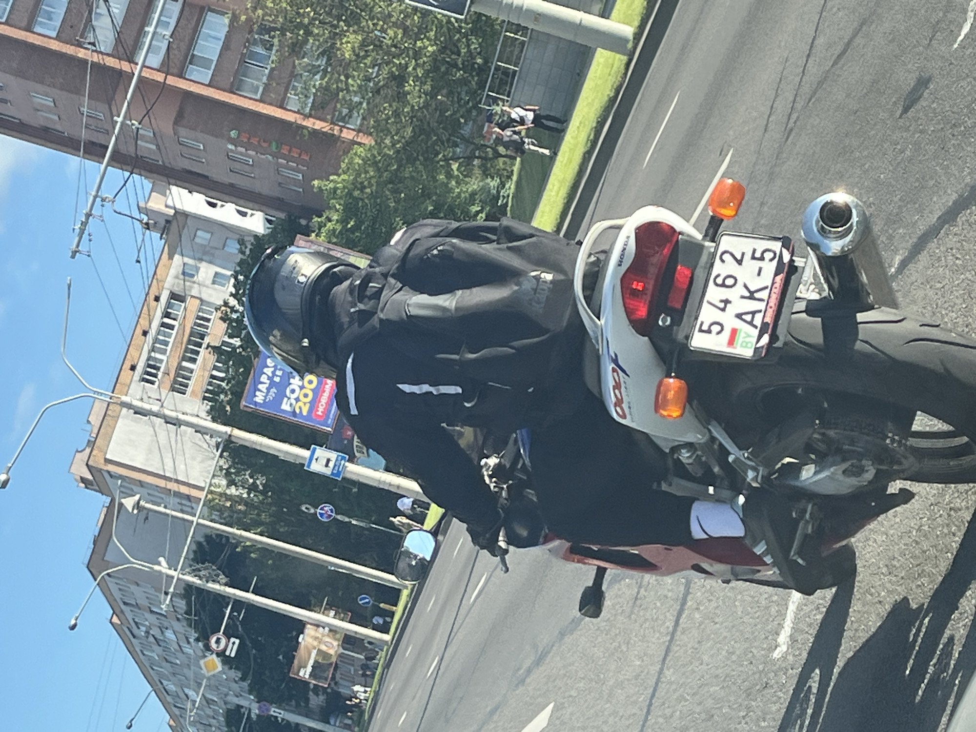 A person is riding a motorcycle down a street in Minsk, Belarus. The rider is wearing a black helmet and black clothing, including a backpack. The motorcycle is red and white and has a license plate that reads "5462 AK-5 BY". The street is lined with buildings and trees, and there are other people walking on the sidewalk. The sky is blue and clear. The image appears to be taken from another vehicle on the street.