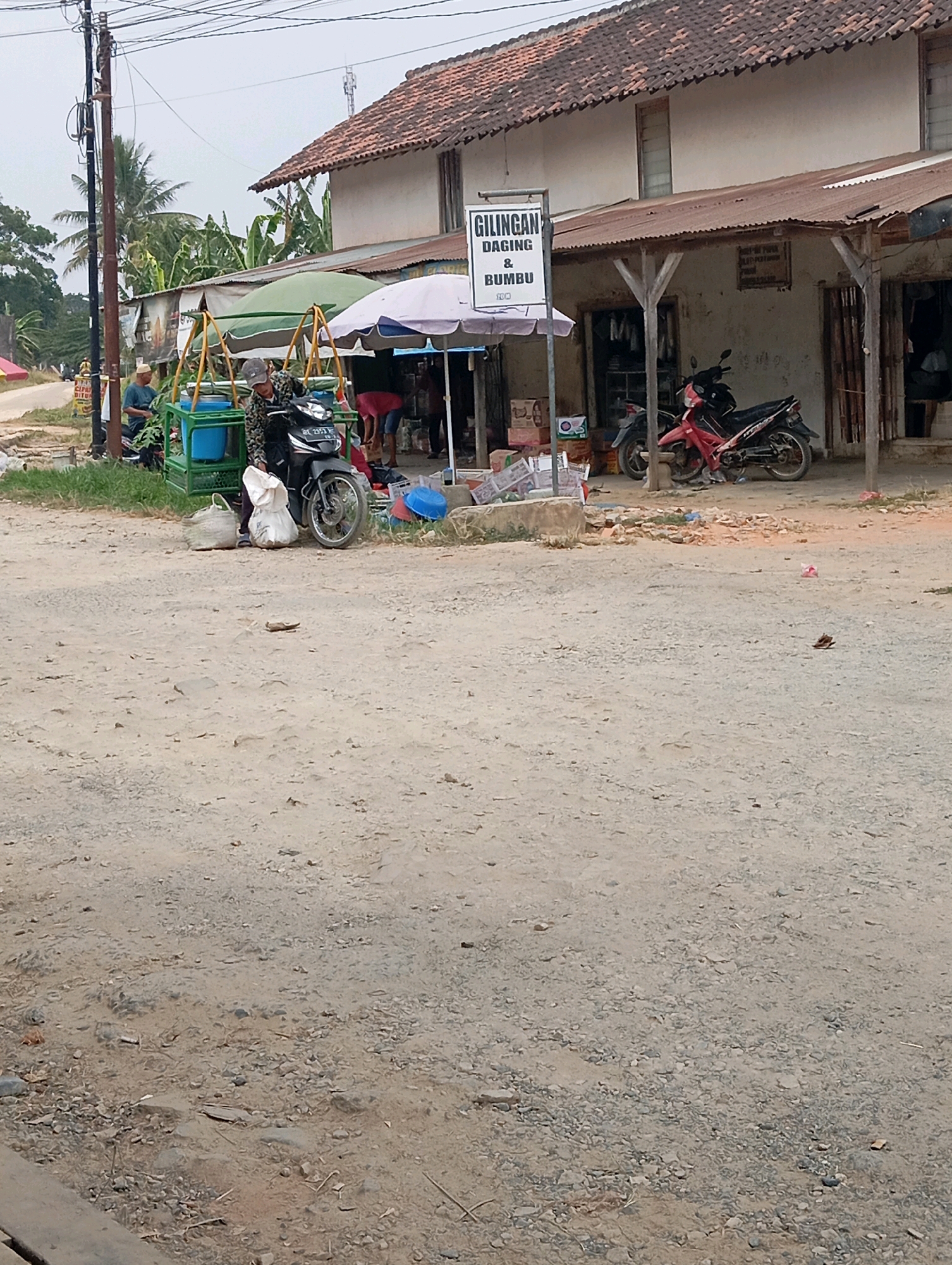 The image depicts a street scene in Leman Benawi, Indonesia, dominated by a dusty, gravelly road. On the right side of the road is a building with a corrugated tin roof, appearing to be a small shop or market stall. A sign above the entrance reads "GILINGAN DAGING & BUMBU" (Meat Grinder & Spices). In front of the shop, two motorcycles are parked, one red and one dark colored. To the left of the shop, a person in a camouflage jacket is seated on the dark motorcycle. Nearby, there is a cart with blue containers and a green frame, and two white sacks are placed on the ground. To the far left, another person is seated on a stool, facing away from the camera. The environment appears to be a rural or semi-urban area, with a power pole and overhead wires visible on the left side of the image. The sky is overcast.