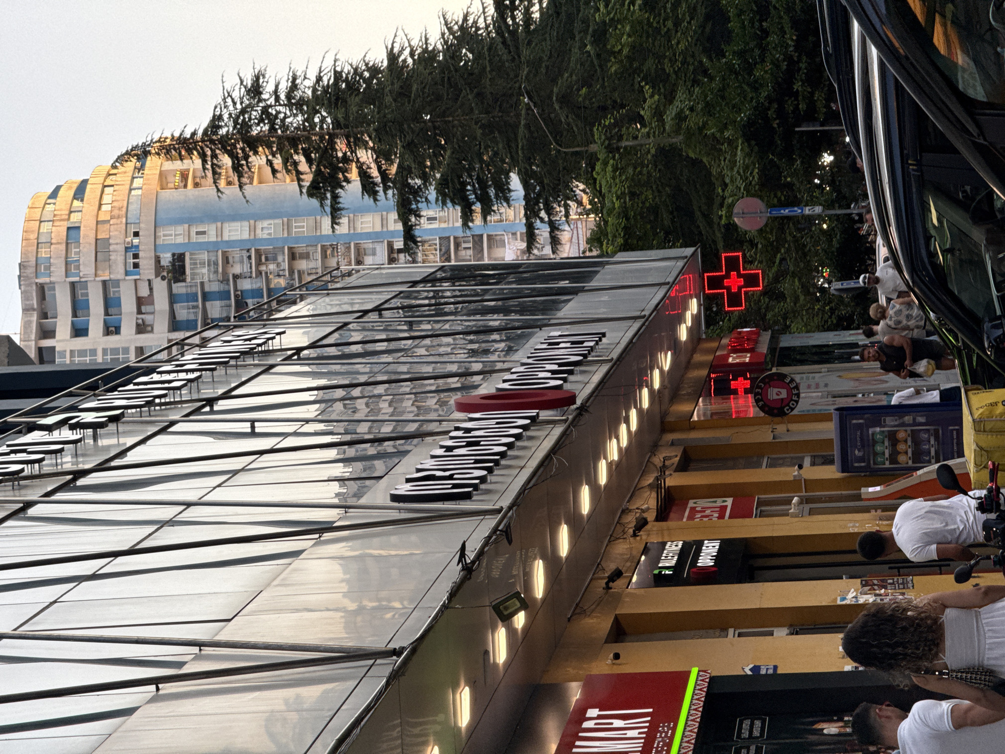 The image depicts a street scene in an urban environment. A modern building with a glass facade dominates the left side of the frame, reflecting the surrounding architecture. To the right, a tall, curved residential building with blue and white accents is visible. A large evergreen tree stands between the two buildings.
In the foreground, several shops line the street. One shop has a prominent red sign with white lettering that reads "MART". Another storefront displays signs for "RULEFITNESS" and "OPPONENT". A bright red, illuminated cross, a common symbol for a pharmacy, is also visible.
Several people are present on the sidewalk, some walking and others standing. A few are dressed in casual clothing, including white t-shirts. A yellow delivery box is placed on a scooter. Cars are parked on the street in the lower right corner of the image. The lighting suggests it is late afternoon or early evening, with warm light illuminating some of the buildings. The sky is a pale, clear color.