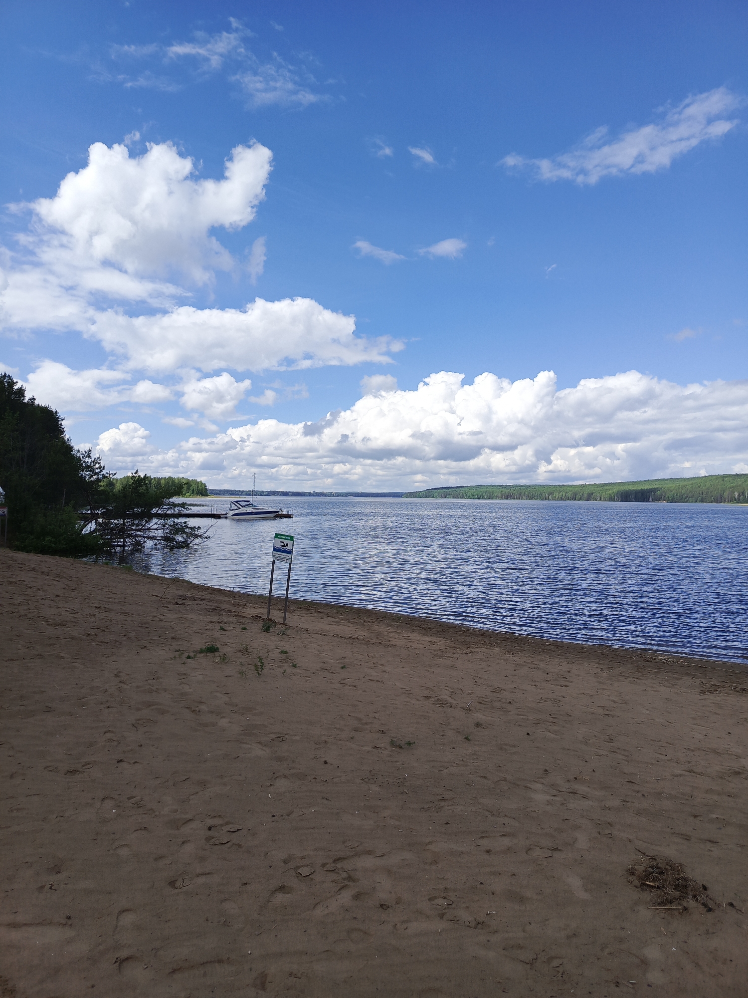 The scene is a daytime view of a lake and beach under a partly cloudy sky. The beach is sandy, and there is a sign post near the shoreline. A small dock with a white boat is visible in the background. Forest covers the far side of the lake, extending along the left side of the scene. The location is Unknown, Unknown. No people are visible.