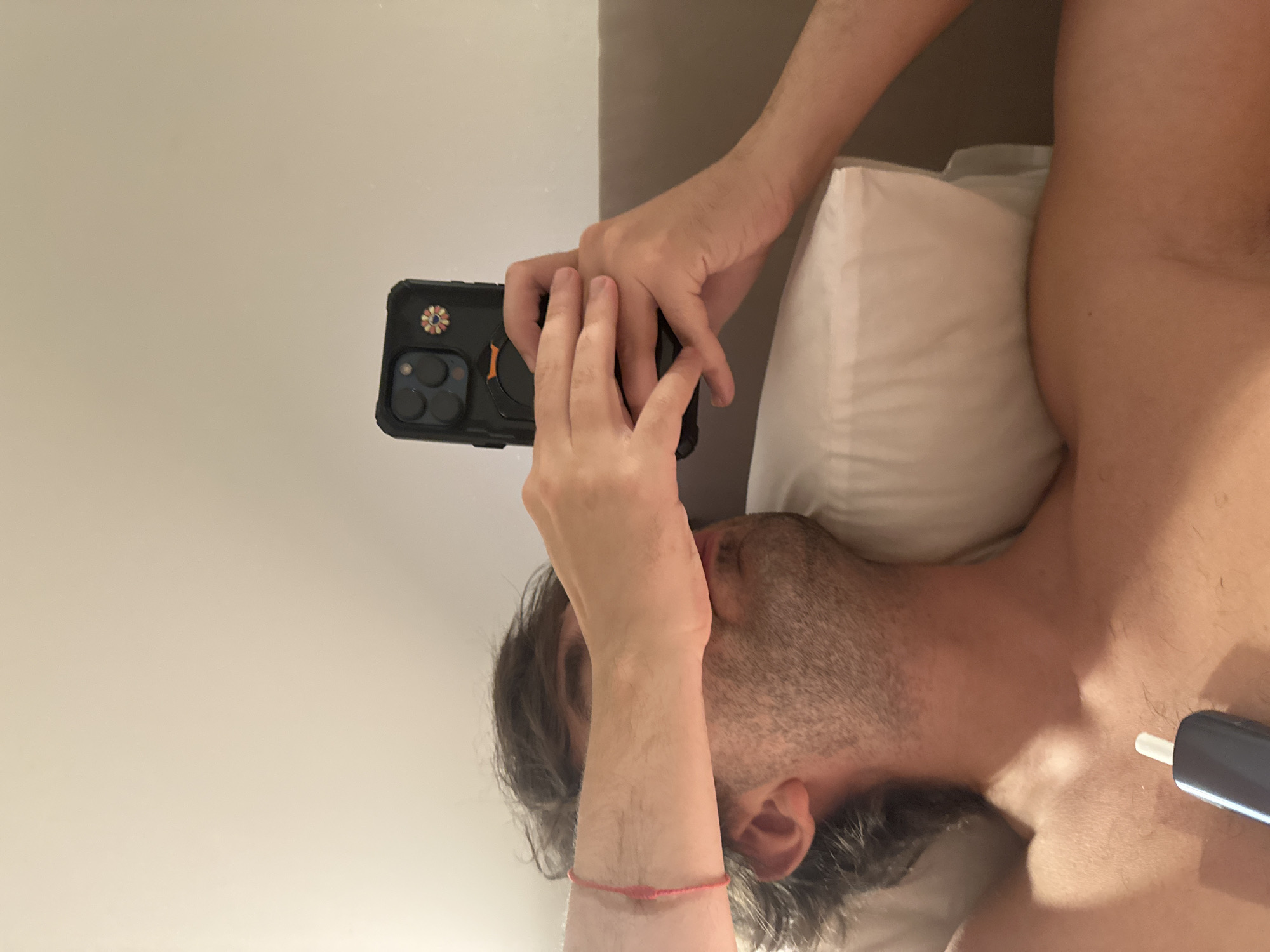 A first-person perspective shot shows a person lying on a pillow in bed, holding a smartphone up to take a selfie. The person's face is mostly obscured by their hand and arm, which are positioned to block the camera. A pink string is tied around the person's wrist. The background is a plain wall, and a headboard is visible behind the pillow. A dark-colored electronic device with a white tip is positioned on the person's chest. The lighting suggests an indoor setting, possibly a bedroom. There are no identifiable landmarks or clues to indicate the location is in Moscow, Russia.