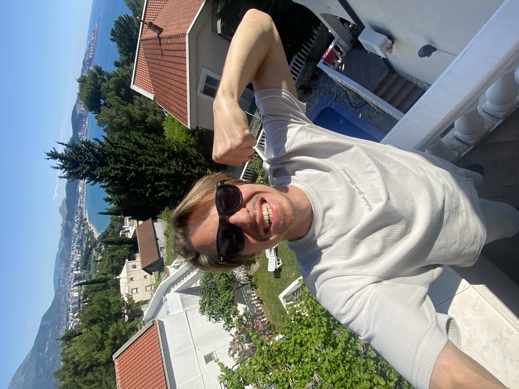A medium shot depicts a man standing on a white balcony, wearing sunglasses and a light-colored t-shirt, holding his arm up in a flexed pose. The background reveals a scenic view of Šušanj, Montenegro, with red-roofed buildings, lush green trees, and the Adriatic Sea stretching into the horizon. A swimming pool is visible in the courtyard below. The man is smiling, indicating a positive emotion or experience.