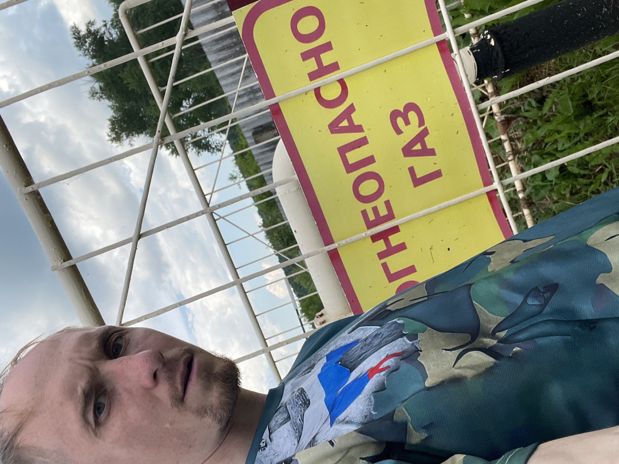 A close-up, low-angle selfie captures a man with light skin, sparse facial hair, and a neutral expression, looking towards the camera. He is positioned in front of a white metal fence. His dark green t-shirt features a camouflage pattern with an emblem on the left chest depicting the Russian flag. Behind the fence, a yellow sign with a pink border is visible, bearing the Cyrillic text "НЕОПАСНО" and "ГАЗ", which translates to "Dangerous" and "Gas". Beyond the sign, a white, cylindrical object, likely a gas tank, and lush green vegetation are present under a cloudy sky. The setting appears to be an outdoor industrial or utility area in Nizhnii Bugrysh, Russia.