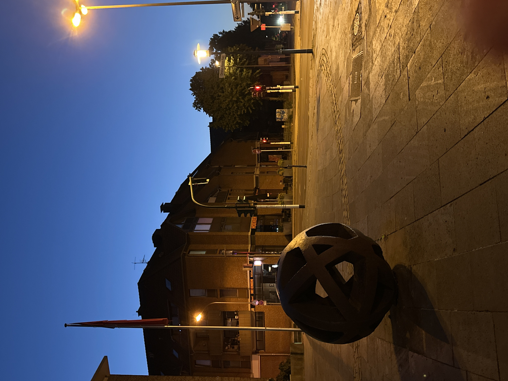 The image depicts a twilight scene in Krefeld, Germany, specifically at the intersection of Hochstraße and Corneliusstraße, near the "Hoenen" establishment, a prominent bakery and café. In the foreground, a large, dark, spherical sculpture with intersecting geometric patterns rests on a paved sidewalk. The sculpture casts a long shadow, indicating the low angle of the artificial street lighting.
The street itself is paved with interlocking stones and slabs, some of which appear damp. A decorative circular pattern is subtly visible in the pavement. To the left of the sculpture, a red flag flies from a tall flagpole, silhouetted against the deep blue twilight sky. Behind the sculpture and flagpole, a multi-story brick building forms the backdrop. This building has several windows and balconies, some illuminated, suggesting interior activity. The building's signage indicates "HOENEN Mühlenbäckerei."
Further into the scene, streetlights illuminate the intersection. Traffic signals are visible, one showing a red light. Trees line the street further down, their foliage dark against the sky. Several street signs are present, including one indicating "Corneliusplatz" and another pointing towards "Schulzentrum." The overall atmosphere is quiet and illuminated by artificial light, characteristic of an urban environment at dusk or dawn. There are no people visible in the image.