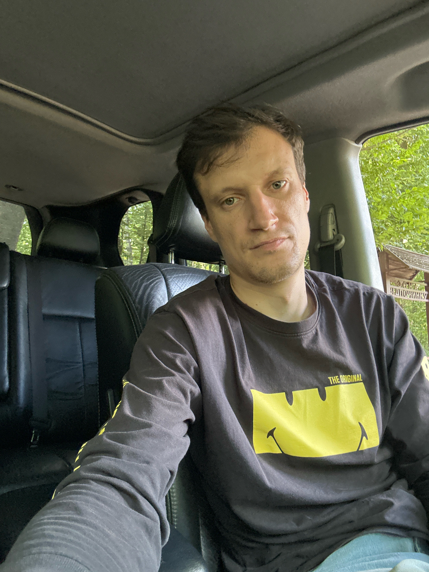 A man with short brown hair and light skin is seated in the driver's seat of a vehicle, facing the camera. He is wearing a dark long-sleeved shirt with a large yellow smiley face graphic and the text "THE ORIGINAL" on it. His left arm is resting on the center console. Behind him, the black leather interior of the car is visible, including the back seats and headrests. Through the windows and sunroof, lush green trees and foliage can be seen, suggesting an outdoor, possibly wooded, environment. A wooden sign with Cyrillic writing is partially visible on the right side of the frame, indicating the location is near Shatava, Ukraine. The overall lighting suggests daylight.