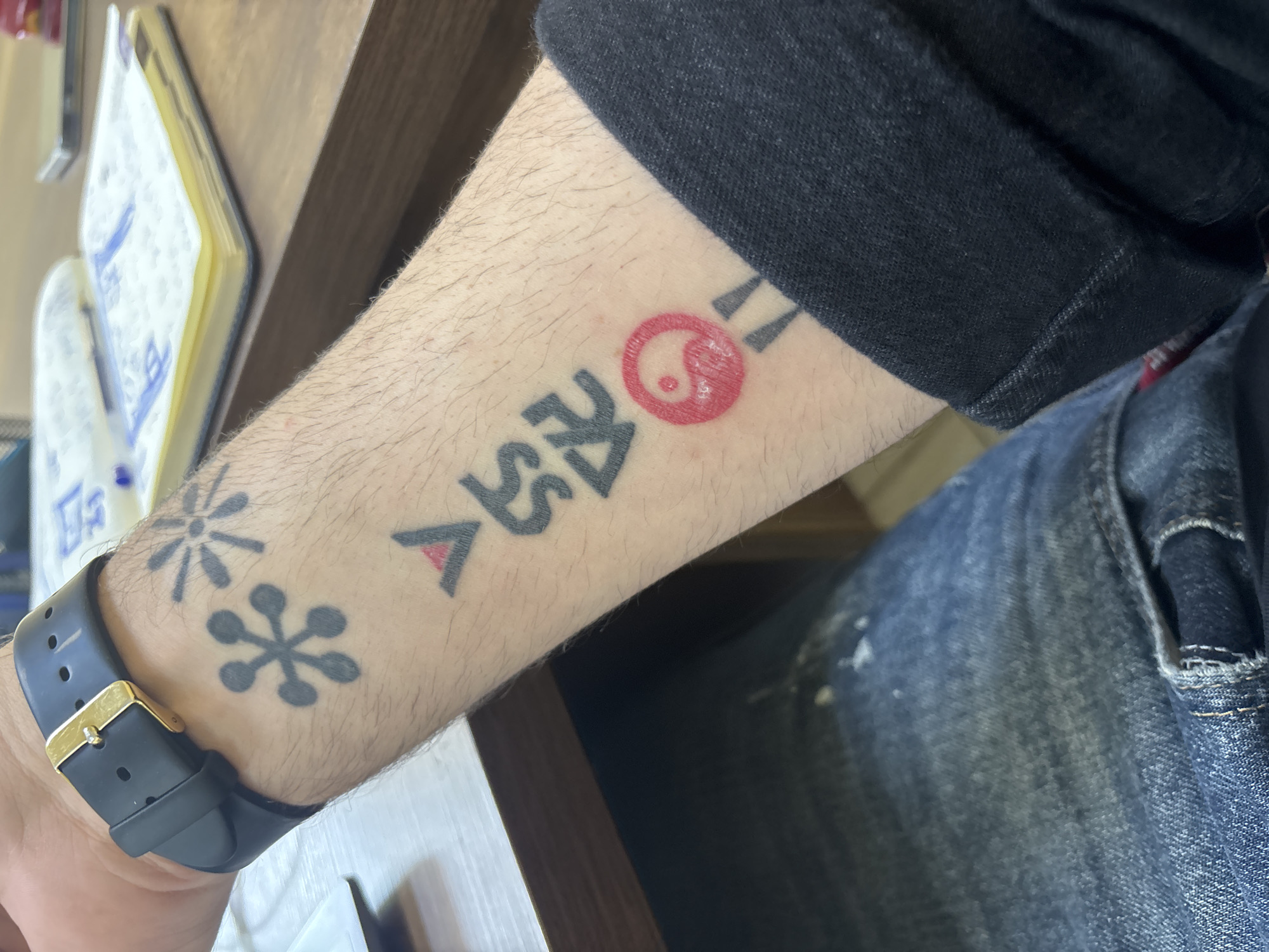 A close-up image depicts a person's forearm and a portion of their body and immediate surroundings. The forearm is slightly angled, revealing several tattoos. The tattoos include stylized floral designs near the wrist, and a combination of symbols and characters further up the arm, one of which resembles a red Yin-Yang symbol with additional markings. The person is wearing a black smartwatch with a gold buckle. Part of a black denim shirt or jacket is visible, as well as blue denim jeans. The forearm rests near a wooden surface, possibly a table or desk, where a notebook is located.
The description does not give any information about this scene being in Tbilisi, Georgia.