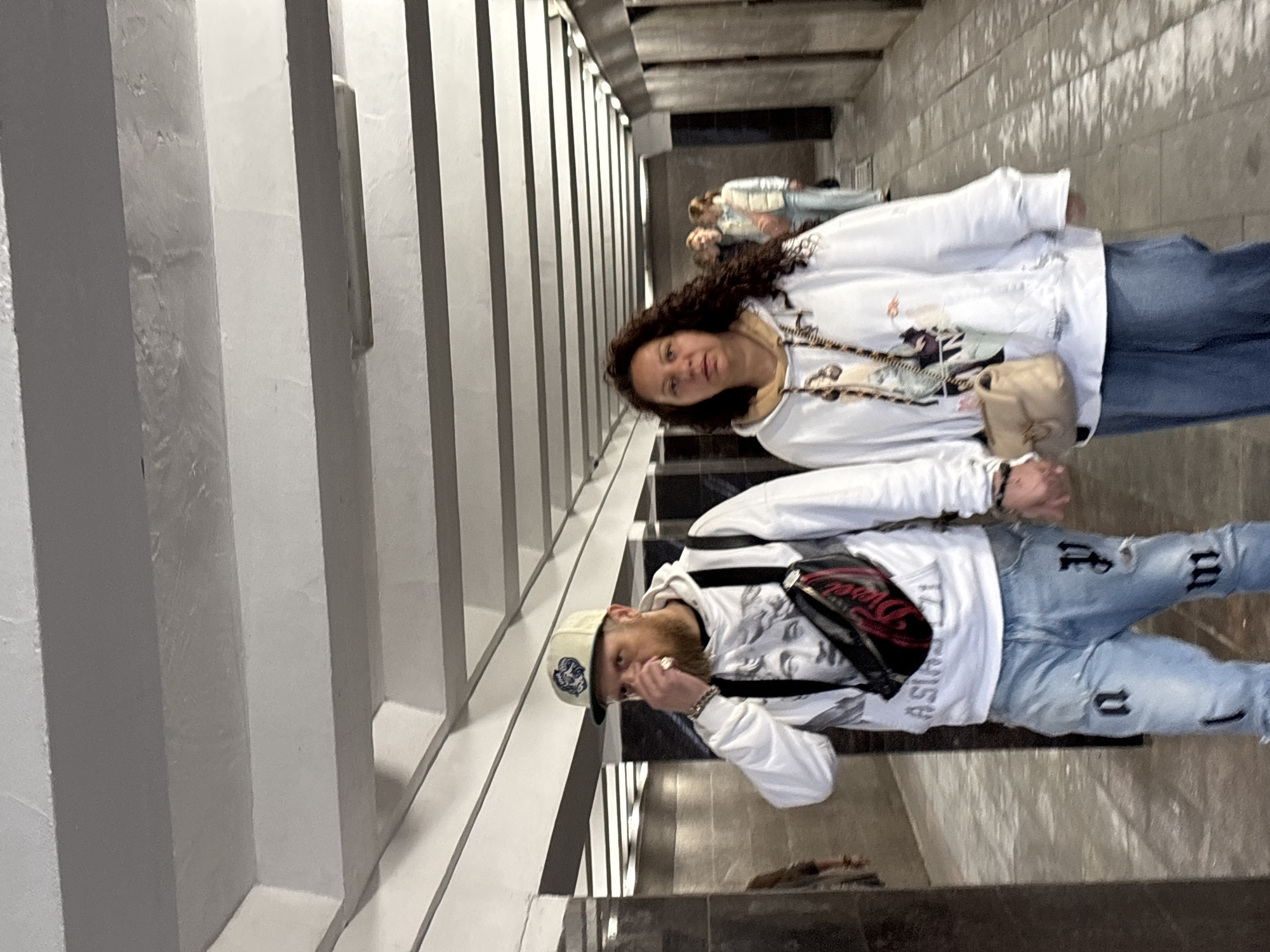 The image depicts a couple walking through an underground passage in Nizhny Novgorod, Russia. The passage is characterized by a white, ribbed ceiling and dark, tiled walls. The couple, a man and a woman, are walking towards the viewer, holding hands. The man is wearing a white hoodie with a graphic print of a bust, blue jeans with black lettering, and a beige baseball cap. He has a beard and is touching his nose. The woman is wearing a white hoodie with a graphic print and blue jeans. She has long, curly brown hair. Behind them, other individuals can be seen walking in the same direction. The passage appears to be an urban underpass or pedestrian tunnel.