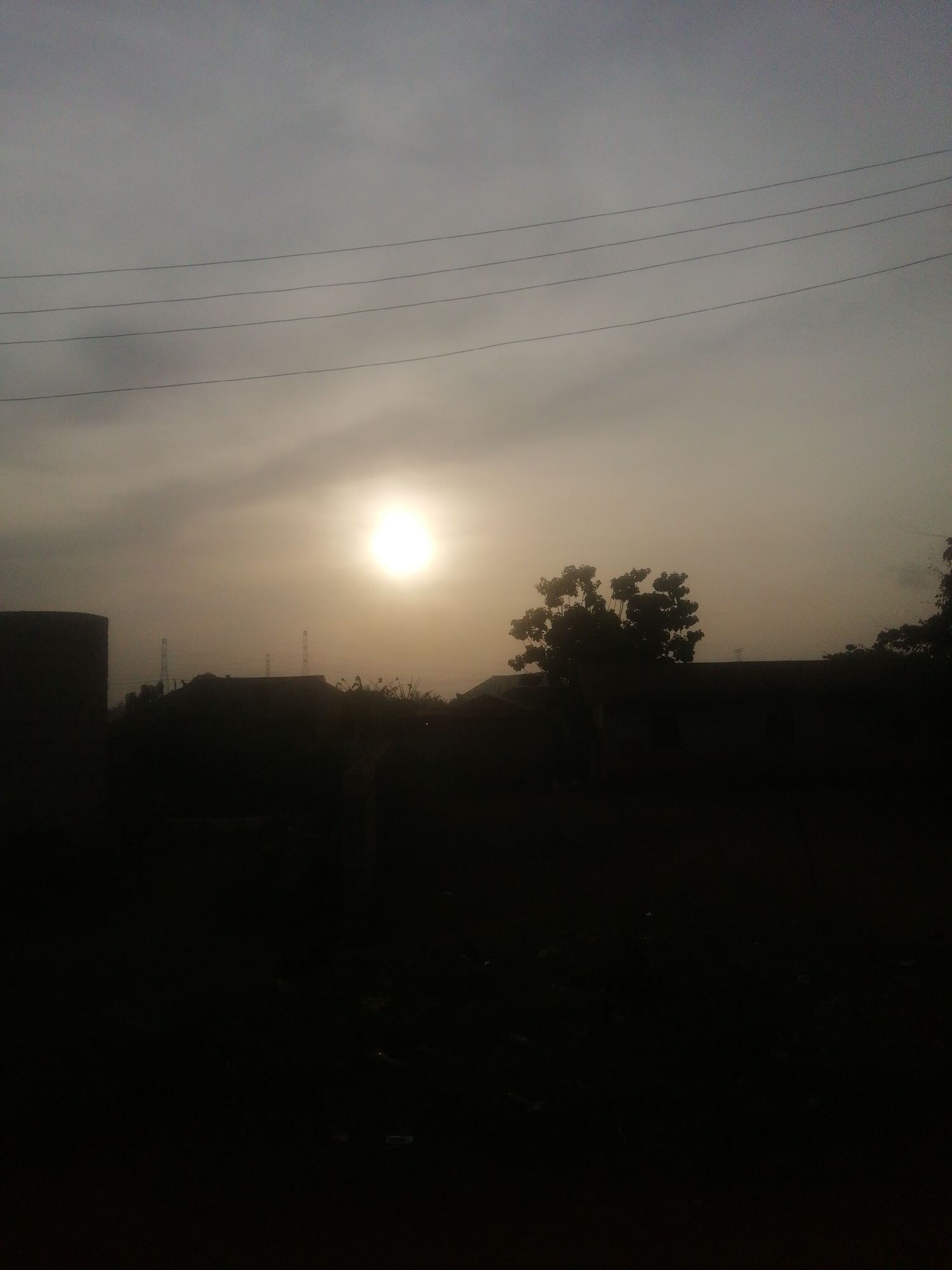The image depicts a silhouetted scene, likely at sunrise or sunset. A bright sun is visible through a hazy sky with overhead power lines. The foreground features dark silhouettes of buildings, trees, and general vegetation, suggesting an urban or semi-urban environment. The exact location is unknown.