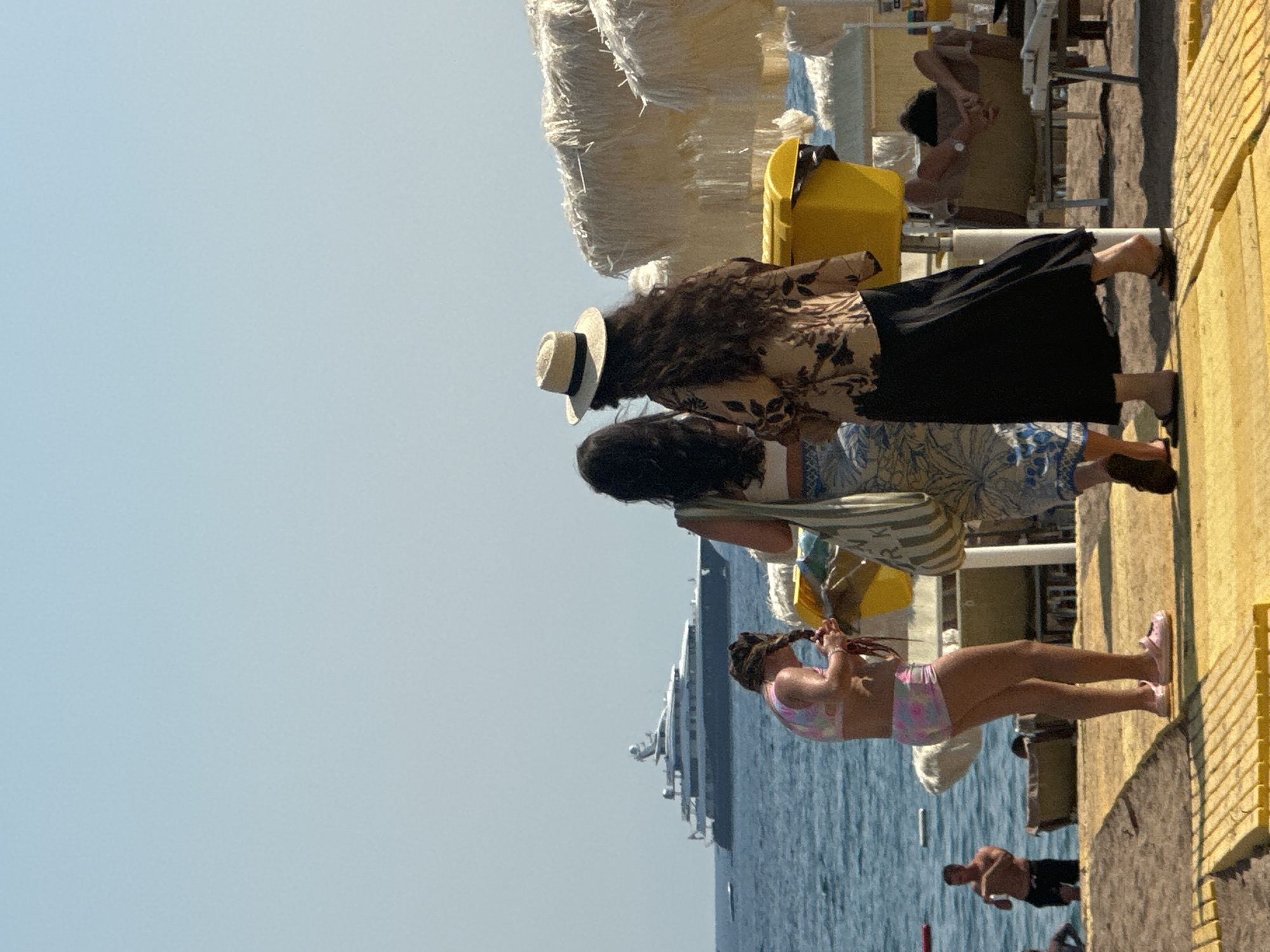 In Boreti, Montenegro, on a sunny day at the beach, three women are standing on a yellow wooden platform. One woman in the foreground, facing away from the camera, wears a pink and white patterned bikini and is braiding her dark hair. To her right, another woman, also facing away, wears a patterned shirt and a long black skirt and a straw hat. The third woman, to her left and partially visible, wears a light-colored top and a patterned skirt. In the background, a large yacht is visible on the calm blue sea. Beach umbrellas with straw fringes and a yellow trash bin are also present. Further back, a man in dark shorts stands near the water. The scene depicts a typical beach day with leisure activities.
