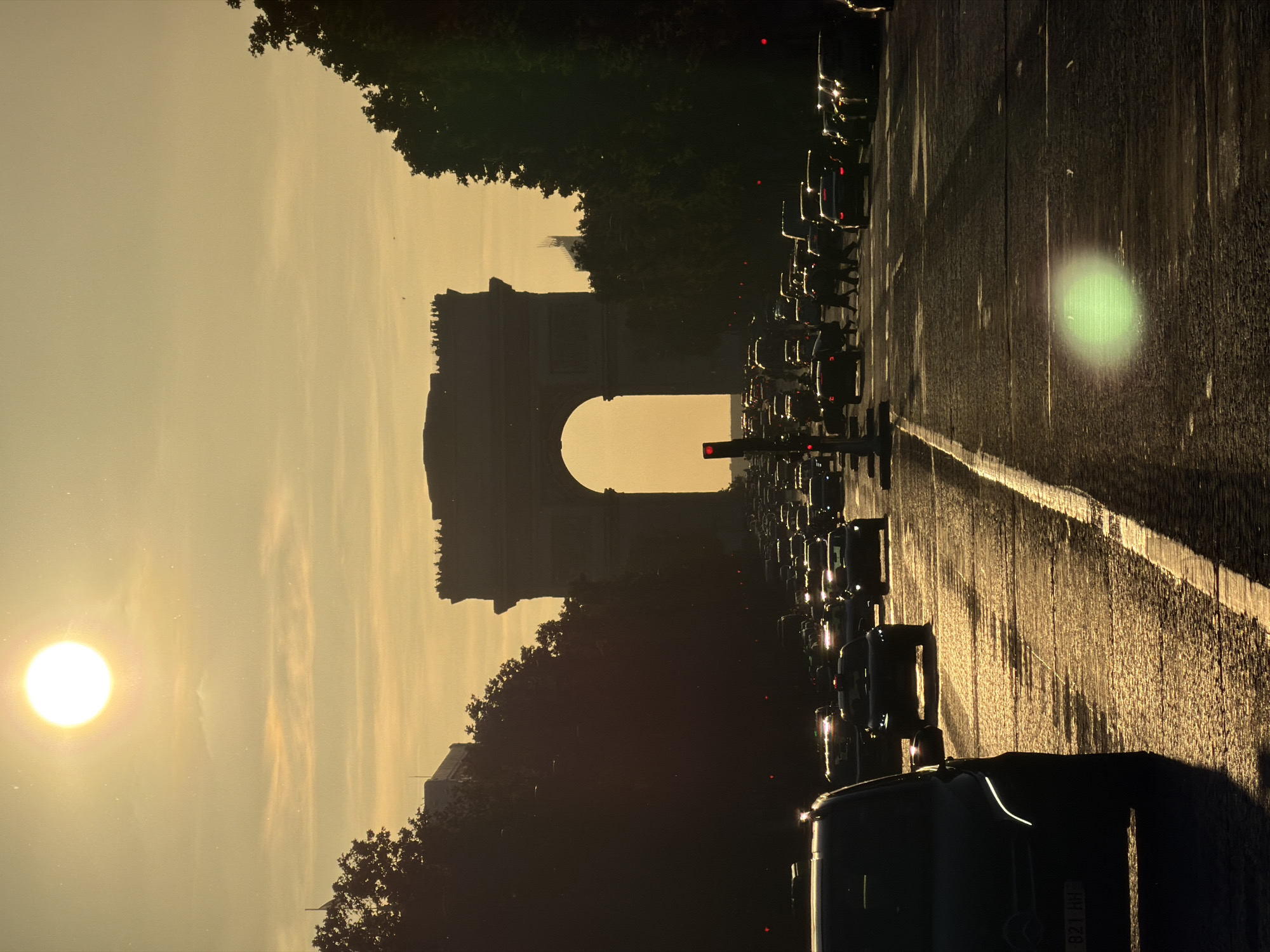 The image shows the Arc de Triomphe in Paris, France, viewed from the Champs-Élysées. Traffic is stopped in the street, a traffic light is visible, and the sun is setting.