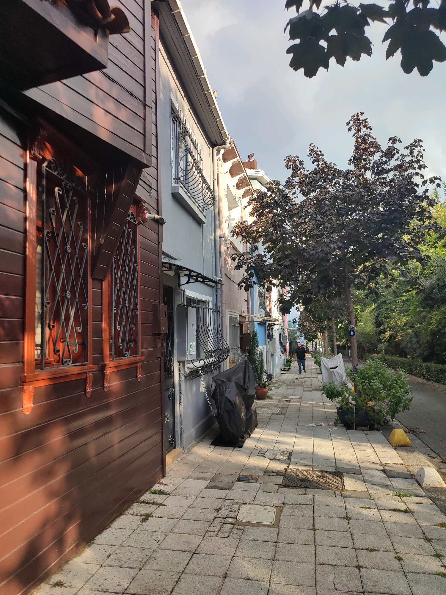 This image shows a street scene in Kadıköy, Turkey, likely a residential area with narrow streets and traditional architecture. In the foreground, a dark brown wooden house features decorative windows with ornate metal grilles. Adjacent to it is a building with a gray facade, also adorned with similar window grilles. A paved sidewalk runs alongside the buildings, leading into the distance where a lone figure, a man wearing a dark t-shirt and blue jeans, is walking away from the camera. The street is lined with trees, one of which has dark purple foliage. Various objects are visible along the sidewalk, including planters, covered items, and utility covers. The environment suggests a temperate climate, with the sun casting long shadows across the pavement. The overall impression is one of a quiet, residential street with a blend of old and new architectural elements.