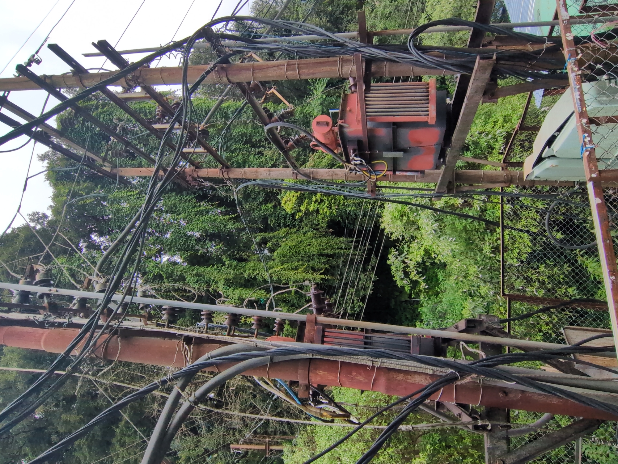 The image captures a dense network of electrical poles and wires in Bhagsu, India. Two prominent, weathered, rust-colored poles dominate the foreground. The pole on the left is a thick, cylindrical structure heavily encrusted with rust and surrounded by a tangle of black and silver electrical cables. Several black cables, some of which appear thick and insulated, are suspended and secured to the pole by various means, including clamps and metal brackets. Interspersed with these are thinner, lighter-colored wires.
The pole on the right is also made of weathered metal, showing significant signs of rust. It supports a complex arrangement of metal arms and braces, onto which more electrical cables are attached. Suspended from these braces is a large, red and black electrical transformer. The transformer has a ribbed metal casing and several input and output wires connected to it.
In the background, lush green foliage, likely trees and vines, forms a dense canopy, suggesting an outdoor, possibly semi-rural or mountainous setting. The foliage creates a stark contrast with the industrial, decaying appearance of the electrical infrastructure. The sky above is overcast and bright, indicating diffused daylight.
A chain-link fence with rusted top railings is visible in the lower portion of the image, running horizontally across the scene, separating the viewer's vantage point from the electrical equipment. In the far lower right, part of a light green electrical enclosure or box can be seen, also behind the fence. The overall impression is one of aging infrastructure set against a vibrant natural backdrop, characteristic of many areas in India where utility services are integrated with the environment. There are no people or animals visible in the scene.