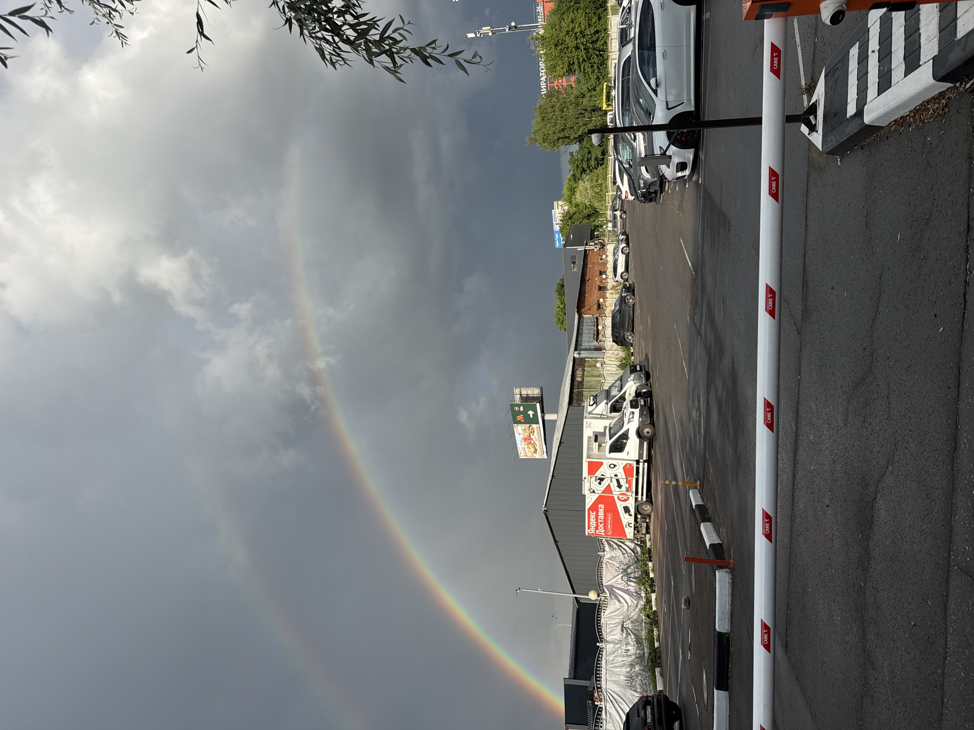 A wide-angle outdoor vertical shot captures a double rainbow against a dramatic, overcast sky. In the foreground, a gray asphalt parking lot is bisected by a white barrier gate with red "CAME" logos repeated along its length. To the right of the barrier, a red and white boom gate mechanism is visible. Further back, in the middle ground, a white delivery truck with a red logo that reads "Яндекс Доставка" (Yandex Delivery) is parked. To the left of the truck, a white tarp covers a structure, and beyond that, a series of parked cars are visible in a lot. In the background, buildings with gray and brick facades stand, one with a large billboard featuring food advertisements. Green trees and a cell tower are also present. The sky transitions from bright white clouds to darker, stormy gray clouds, with the double rainbow arcing prominently across the scene. The overall impression is a post-rain or pre-storm atmosphere.