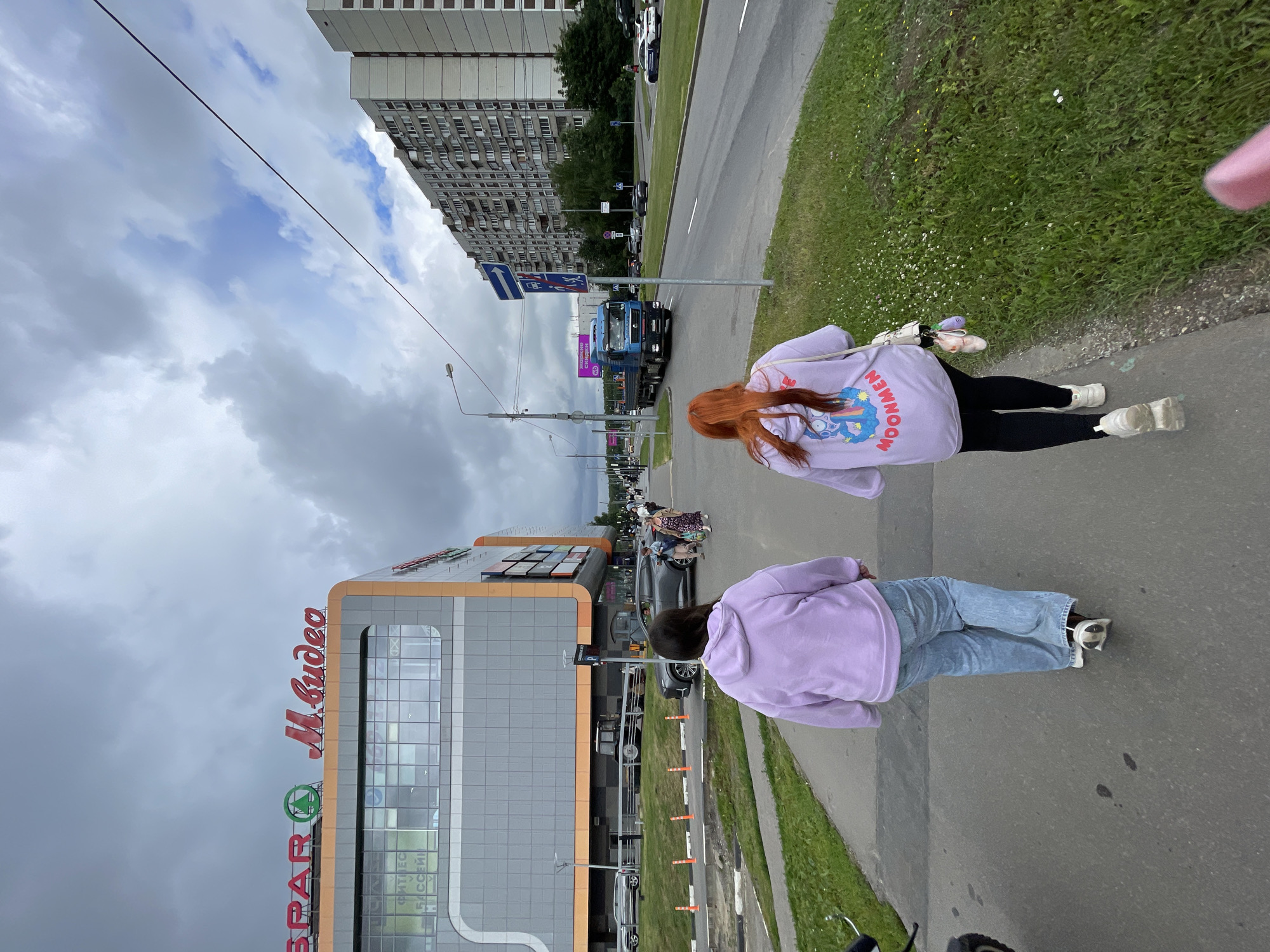 The image captures a street scene in Moscow, Russia, on an overcast day. In the foreground, two young women walk away from the camera on a paved sidewalk bordering a grassy area. The woman on the left wears a light purple hooded sweatshirt and blue jeans, while the woman on the right has red hair and wears a similar purple sweatshirt with black leggings. Both women are walking.
To their left, a large, modern commercial building dominates the left side of the frame. It features a gray facade with large glass windows and prominent signage that includes "SPAR" with a green logo and "М.видео" in red cursive. The building appears to be a shopping center, with an orange trim along its upper edge. Several cars are parked or driving in front of the building.
To the right of the sidewalk, a multi-lane road stretches into the distance, with a blue flatbed truck in motion. Buildings typical of Soviet-era apartment blocks line the far side of the road, appearing tall and densely constructed. Traffic signs and streetlights are visible along the roadside. The overall environment suggests an urban setting within Moscow, likely in a commercial or residential district. The weather appears to be cloudy, with a mix of gray clouds and patches of lighter sky.