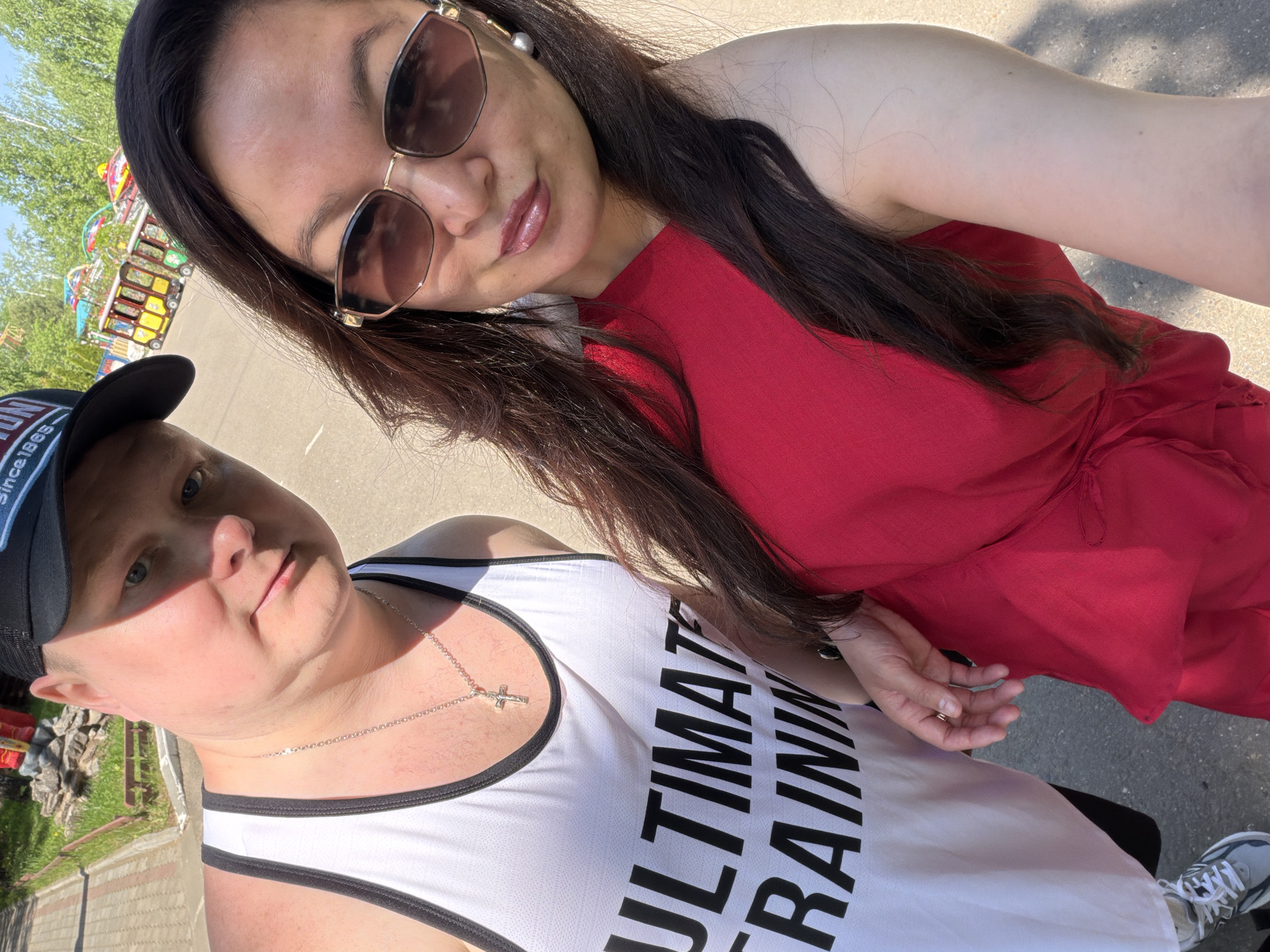 A medium shot of two people, one wearing a black baseball cap and a white tank top with the words "ULTIMATE TRAINING" in black letters, and the other wearing sunglasses and a red dress, on a paved surface, possibly a sidewalk, in an outdoor setting. A small amusement park ride is visible in the background, indicating the location may be a park or recreational area.