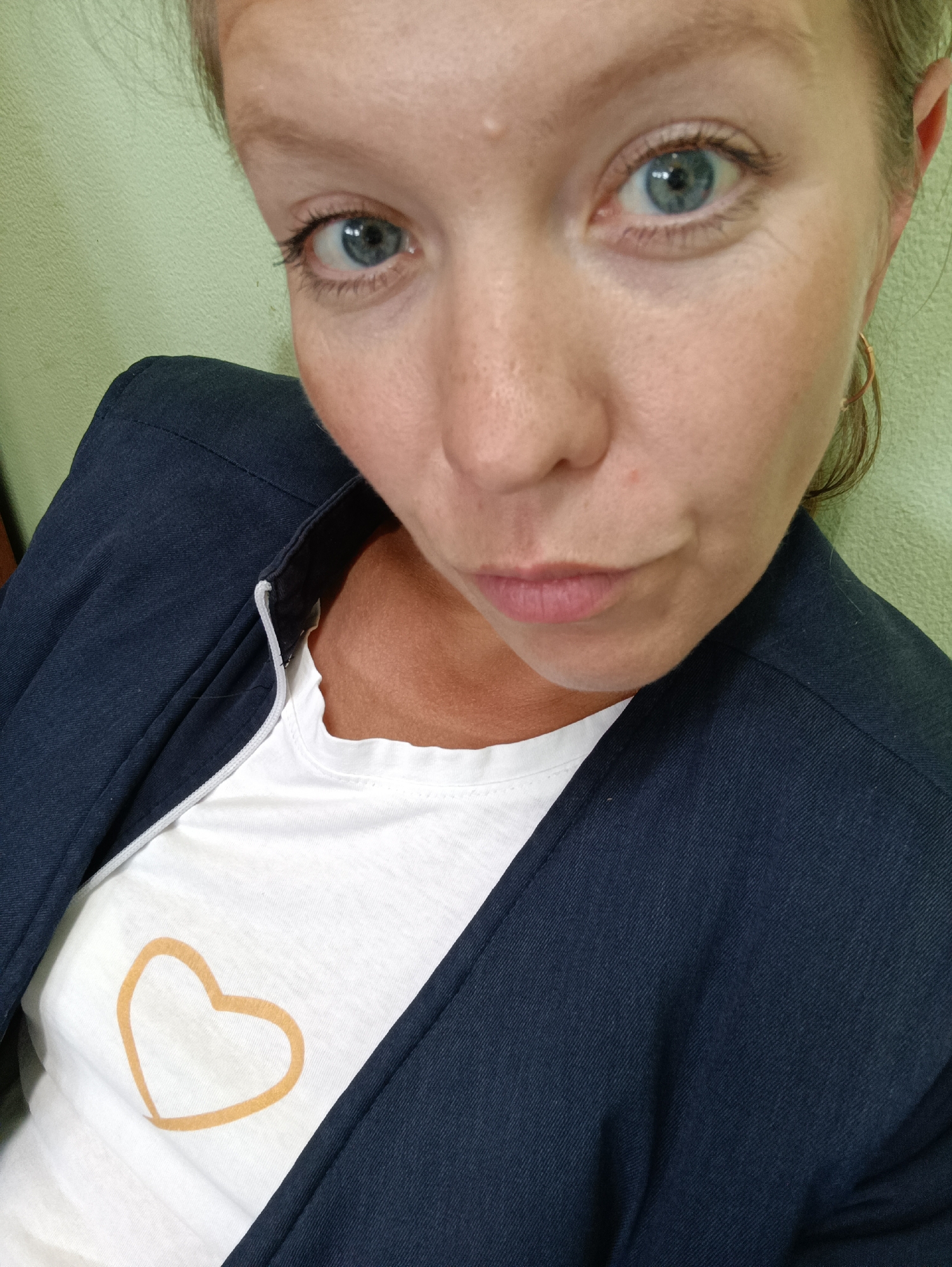 A close-up, selfie-style shot of a fair-skinned woman's face and upper body is presented against a light green textured wall. The woman has blue-grey eyes, light brown eyebrows, and visible eyelashes. Her lips are closed, and a slight pout is visible. She is wearing a dark blue blazer over a white t-shirt adorned with a light orange heart graphic. A gold hoop earring is visible on her right ear. The lighting is somewhat flat, and there are no discernible geographic landmarks or identifying features of Chaikovsky, Russia, visible in the image.