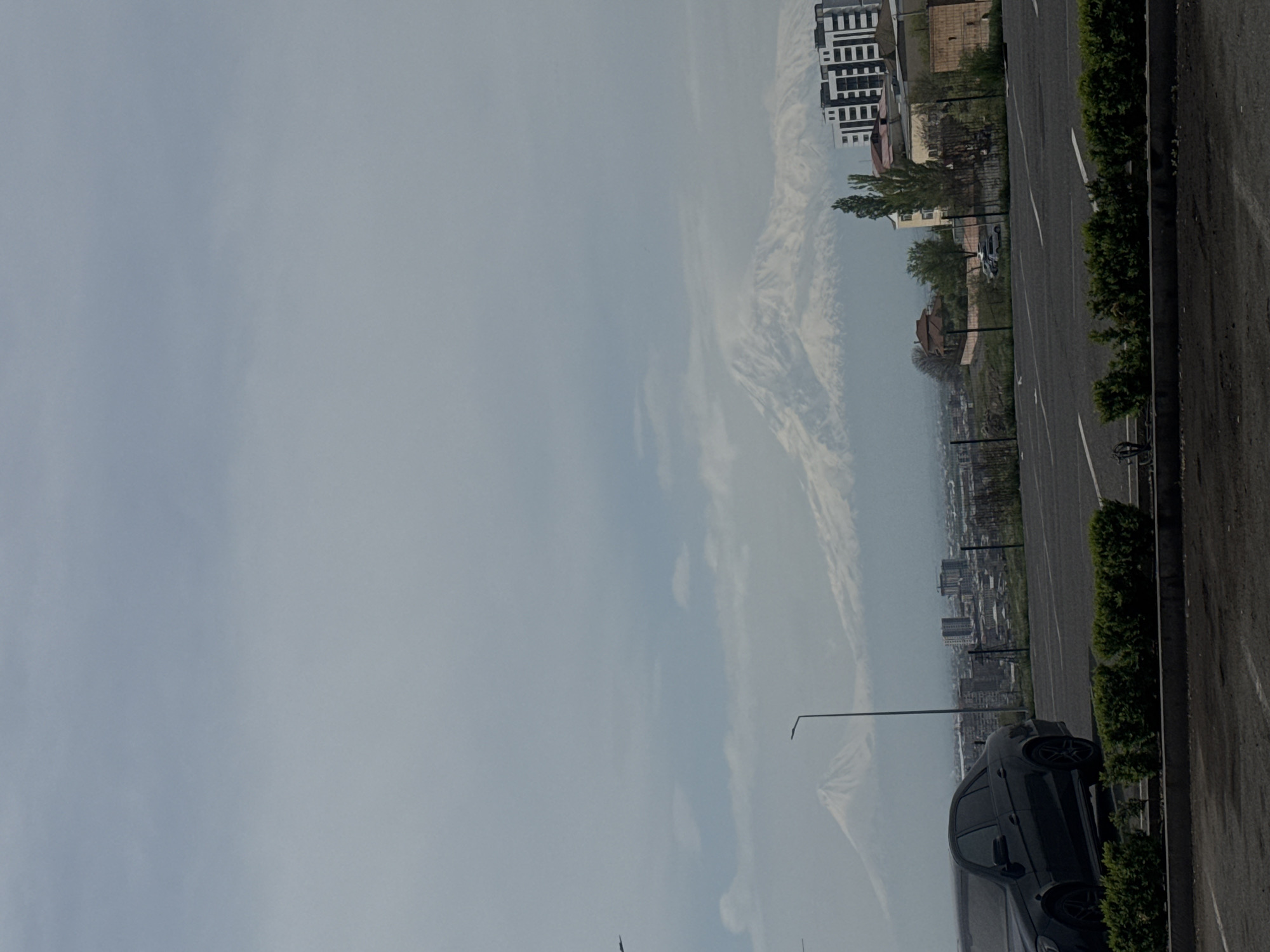 The image presents an urban landscape with a clear sky partially obscured by atmospheric conditions, possibly light clouds or haze, hinting at Mount Ararat in the distance. The lower portion features a dark-colored car parked alongside a road with a narrow border of shrubs. Beyond the road, a cityscape stretches into the distance, characterized by numerous buildings. In the upper portion, there are high-rise apartment buildings, and to the right, a parking lot. The setting appears to be Yerevan, Armenia.