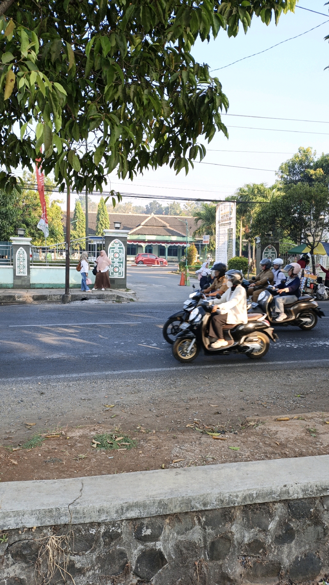 The image captures a street scene in Kuwasen, Indonesia, on a bright, sunny day. In the foreground, a road with a gray concrete curb and a stone wall runs horizontally across the bottom of the frame. The road is partly covered with gravel and dry leaves. On the right side of the road, a concrete barrier with a stone foundation is visible.
The main focus of the image is a group of people riding motorcycles on the asphalt road. Three motorcycles are clearly visible, with riders wearing helmets. The rider of the first motorcycle, in the center of the frame, is a woman wearing a light-colored jacket and a face mask. She is looking ahead while riding. Behind her, two other motorcycles are also moving. The second motorcycle has two people on it, both wearing helmets. The third motorcycle also has two people, with the rear passenger holding a bag.
On the left side of the road, a white building with a green roof and red trim is visible in the background. In front of the building, there is a fence with decorative white pillars. Two women are standing near the fence, conversing. One is wearing a hijab and a long dress, and the other is wearing a hijab and a white shirt with jeans. A red car is parked in front of the building. A flag on a pole is also visible.
Tall trees with green leaves overhang the scene from the top of the frame, casting shadows on the road. The sky above is clear and blue. The overall atmosphere suggests a typical day in a suburban or semi-urban area of Indonesia.