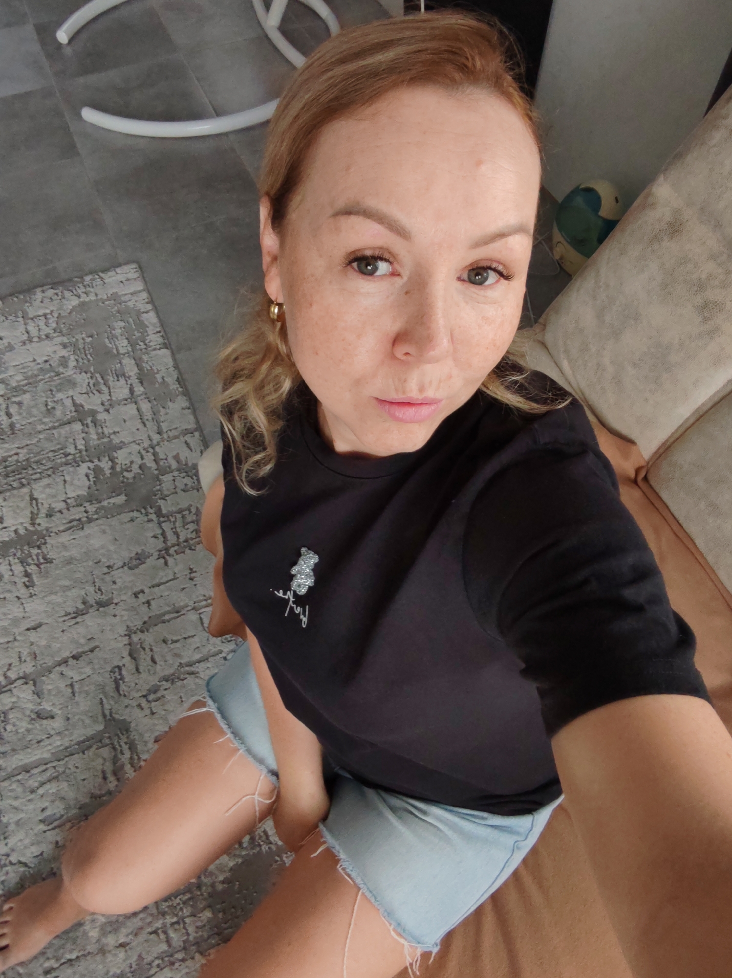 A woman with reddish-blonde hair sits on a brown sofa, wearing a black T-shirt with a small teddy bear design and light blue denim shorts. She looks directly at the camera. The flooring appears to be gray tile with a rug partially visible. A white chair is also partially visible in the background. A blue and white object sits between the woman and the sofa pillows. The woman is barefoot. It is not possible to determine if this photo was taken in Chaikovsky, Russia.