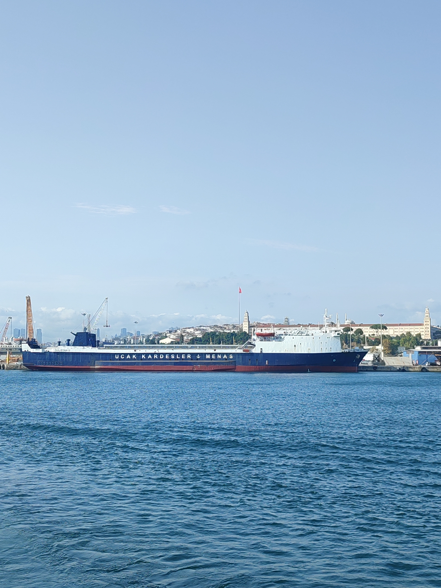 In Istanbul, Turkey, a broad expanse of blue water is in the foreground. Beyond the water, a large navy blue cargo ship is docked. The ship has red trim along its hull. The name of the ship, "UÇAK KARDEŞLER MERSIN," is printed in white lettering on its side. In the background, a city skyline rises, featuring numerous buildings, some with distinct architectural styles including domed structures and towers. A Turkish flag flies from a tall flagpole. Cranes are visible on the left side of the image, suggesting the presence of a port or shipyard. The sky above is a clear, pale blue with a few wispy clouds. The overall impression is one of a busy port scene on a clear day in Istanbul.